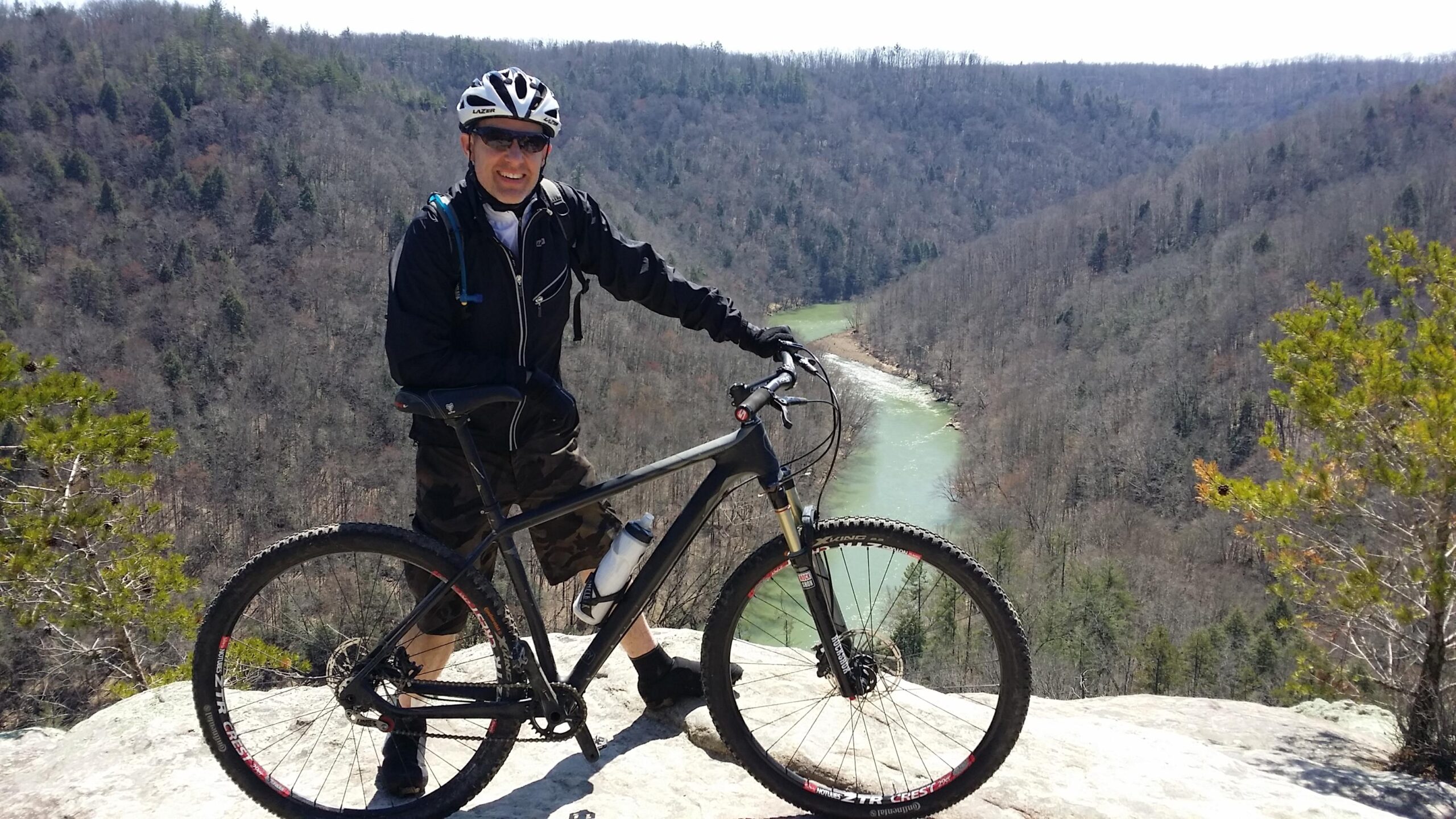 A cyclist poses with a mountain bike on a rocky overlook, showcasing a scenic view of a winding river surrounded by wooded hills. The setting is bright and sunny, with trees in the background and the cyclist wearing a helmet and sunglasses for safety. Big South Fork mountain bike trail.