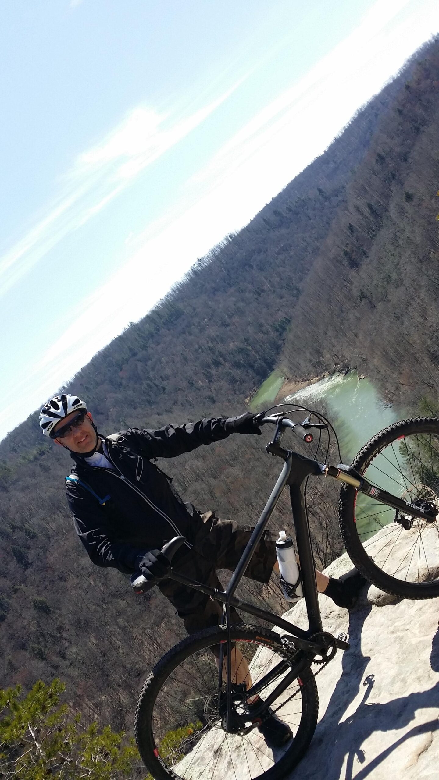 A mountain biker standing on a rocky ledge, wearing a helmet, sunglasses, and a black jacket, with a scenic view of a river winding through a wooded landscape in the background. The sky is clear with a few clouds. Big South Fork mountain bike trail.