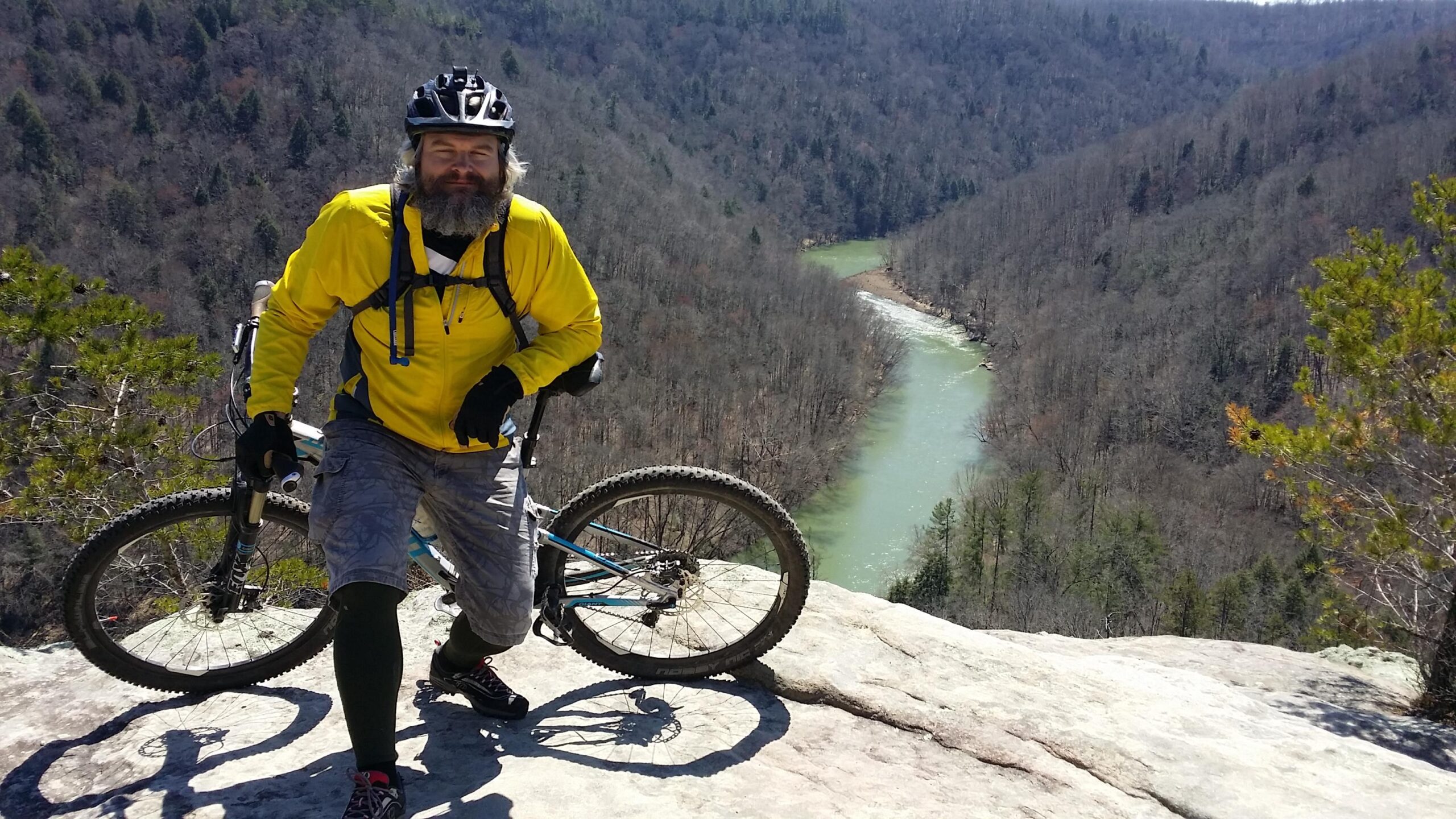 A mountain biker in a yellow jacket poses on a rocky ledge overlooking a winding river surrounded by trees. The landscape is hilly and lush, showcasing a mix of deciduous trees without leaves. The biker is leaning against his bike, wearing a helmet and biking gear, with a cheerful expression on his face. Big South Fork mountain bike trail.