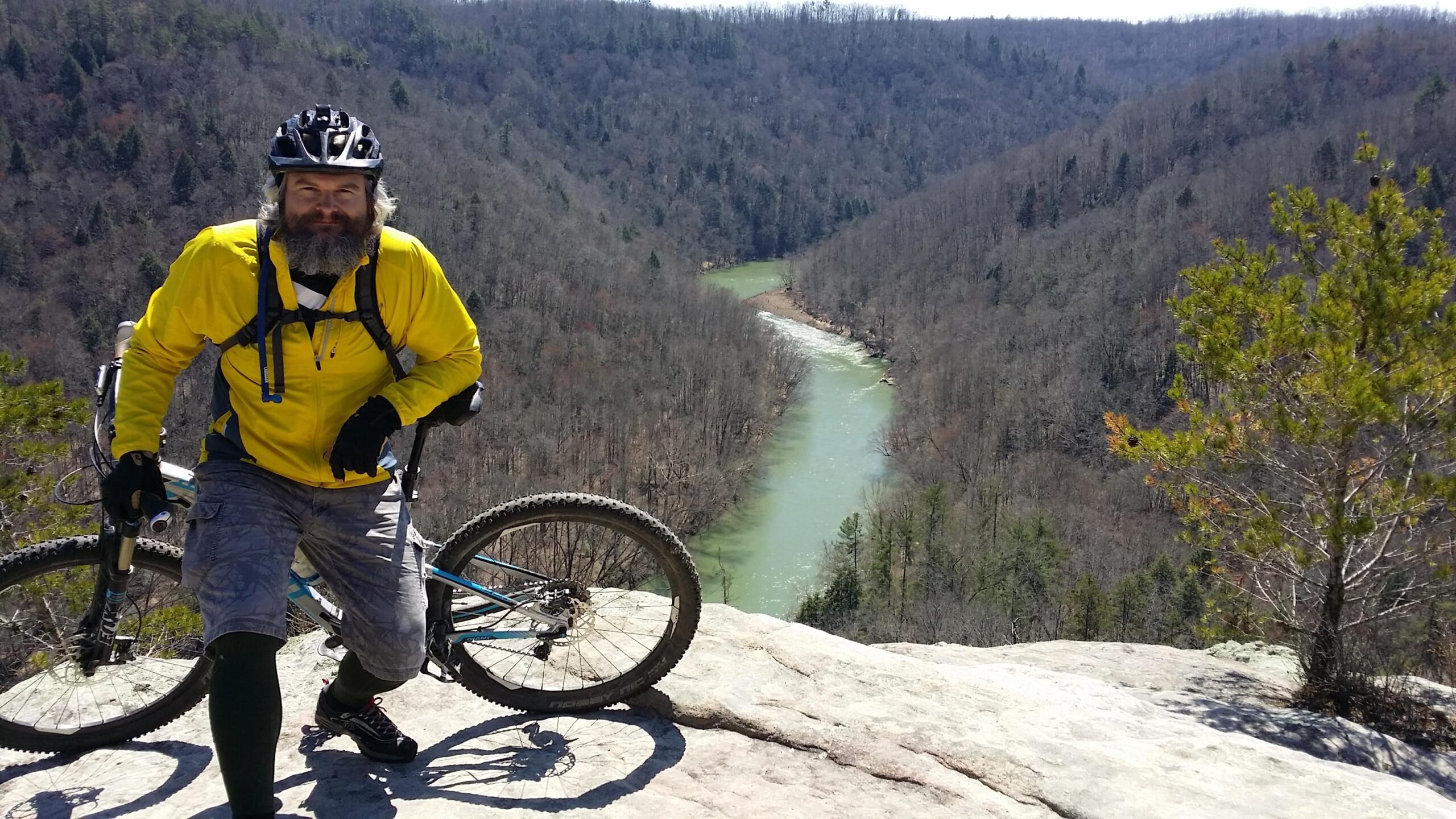 A man with a gray beard dressed in a bright yellow jacket, gray shorts, and gloves stands next to his mountain bike on a rock outcrop, overlooking a winding river surrounded by wooded hills in the background. The scene captures a sunny day in a natural landscape. Big South Fork mountain bike trail.