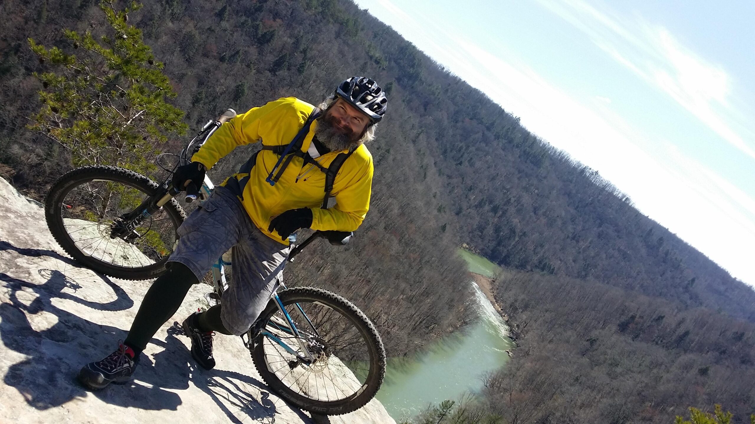 A mountain biker in a bright yellow jacket poses on a rocky outcrop overlooking a winding river and forested landscape, with a clear sky in the background. The biker has a beard and is wearing a helmet, gloves, and shorts, showcasing a spirit of adventure in a scenic outdoor setting. Big South Fork mountain bike trail.