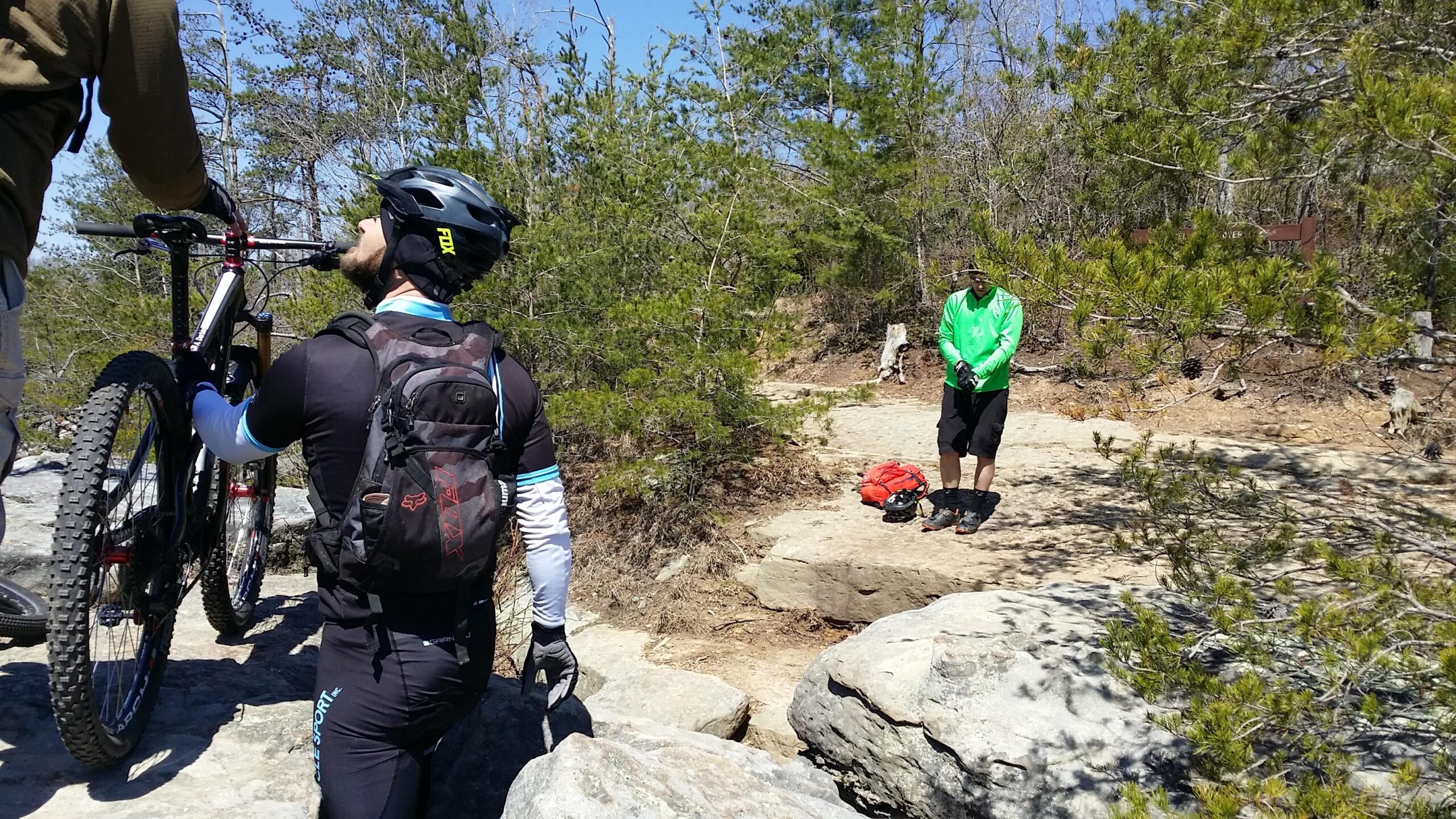 Two mountain bikers adjust their bikes on a rocky trail surrounded by trees on a clear day. One biker is holding a bike while looking up, and the other is wearing a bright green shirt and is standing nearby with a backpack. Big South Fork mountain bike trail.