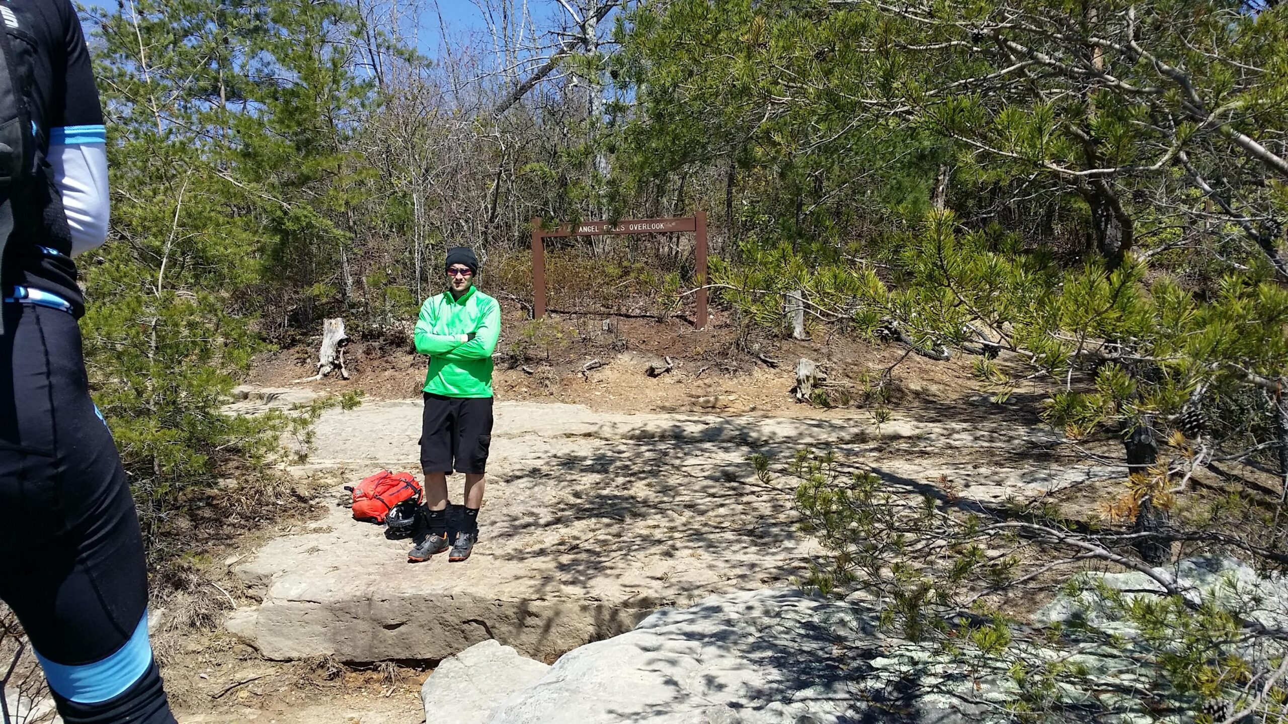 A person in a bright green jacket and black shorts stands on a rocky surface in a wooded area, with an orange backpack nearby. In the background, there is a sign that reads "Angel Falls Overlook." The scene is sunny, with trees and a clear blue sky visible. Big South Fork mountain bike trail.