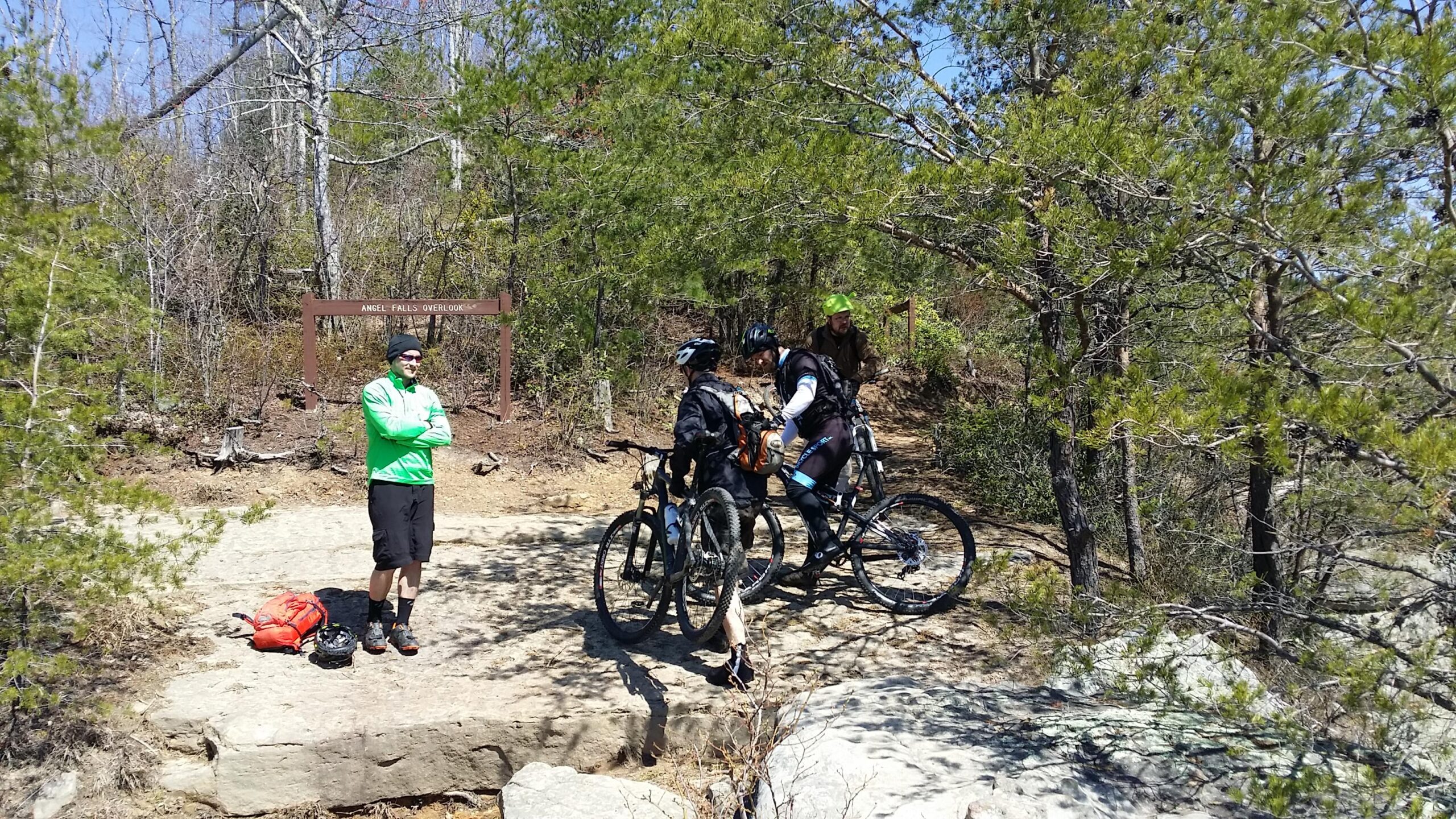 Three mountain bikers preparing for a ride at Angel Falls Overlook, surrounded by trees and rocky terrain. One person stands off to the side in a green jacket, while two others adjust their bikes. A backpack and helmet are placed on the ground nearby. Big South Fork mountain bike trail.