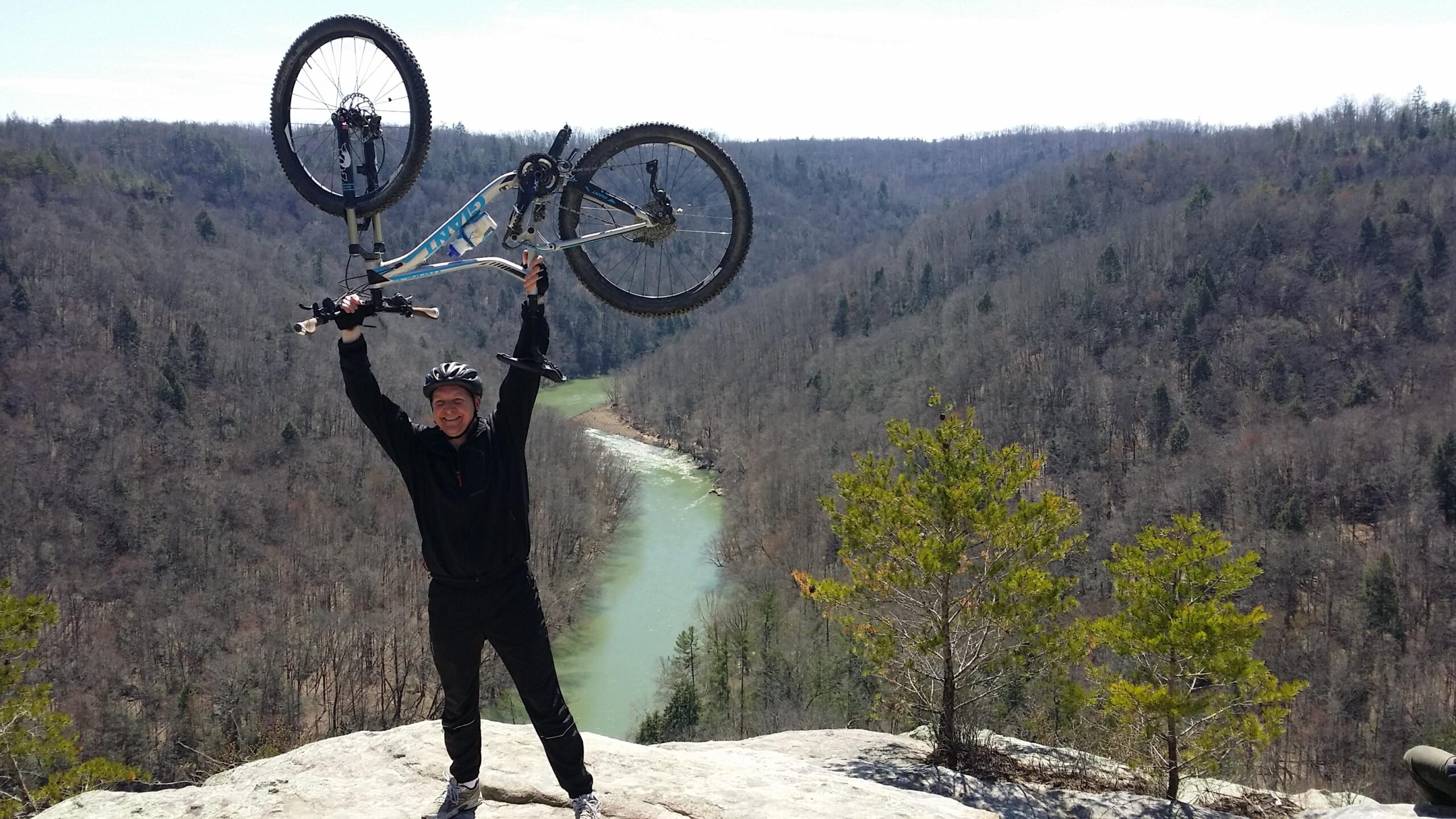 A smiling individual in a black helmet and jacket raises a mountain bike above their head while standing on a rocky ledge overlooking a winding river and forested valley. The scene captures a sense of achievement and the beauty of nature on a clear day. Big South Fork mountain bike trail.