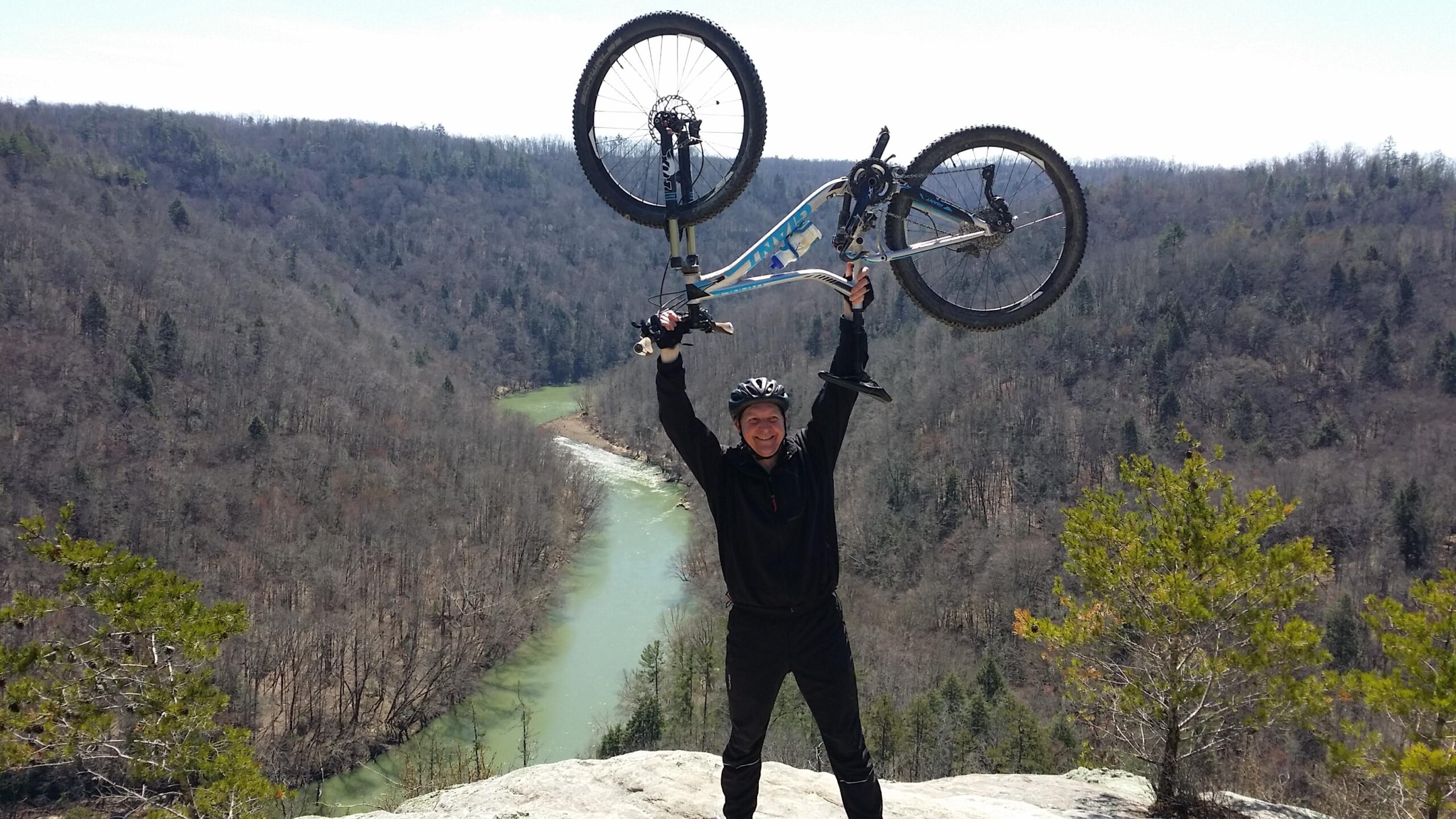 A mountain biker wearing a helmet raises their bicycle triumphantly above their head on a rocky overlook, with a scenic view of a winding river and wooded hills in the background. Big South Fork mountain bike trail.