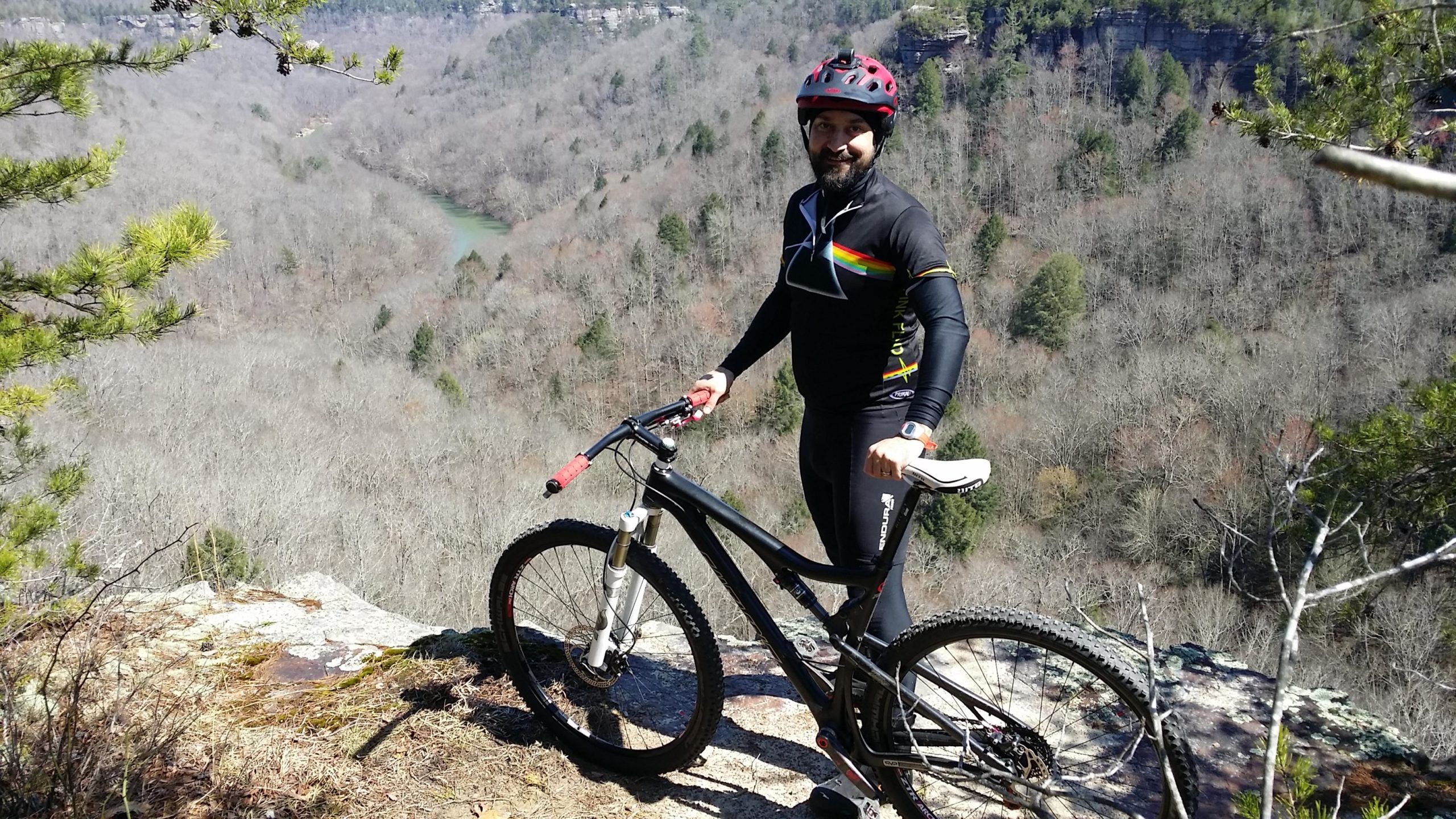A mountain biker stands on a rocky ledge overlooking a valley with a winding river. The cyclist is wearing a black jersey with colorful stripes, a red helmet, and holding onto a mountain bike with thick tires. The background features trees with bare branches, characteristic of a late winter landscape, under a clear blue sky. Big South Fork mountain bike trail.