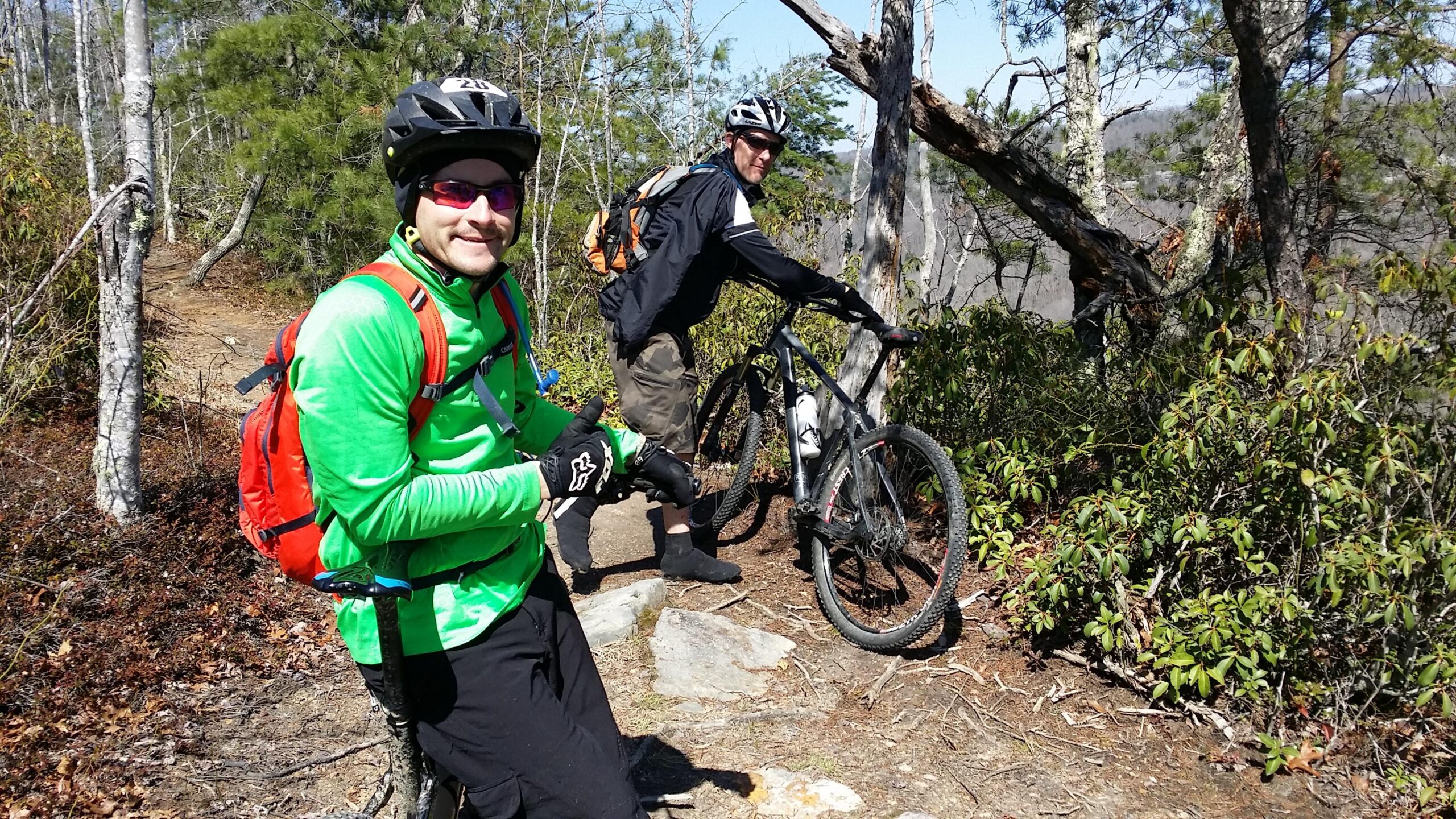Two mountain bikers pause on a forest trail, one giving a thumbs up with a smile while wearing a bright green long-sleeve shirt, helmet, and gloves. The other biker stands next to a black mountain bike, adjusting gear, dressed in a black jacket and shorts. The surrounding landscape features trees and shrubs under a clear blue sky. Big South Fork mountain bike trail.
