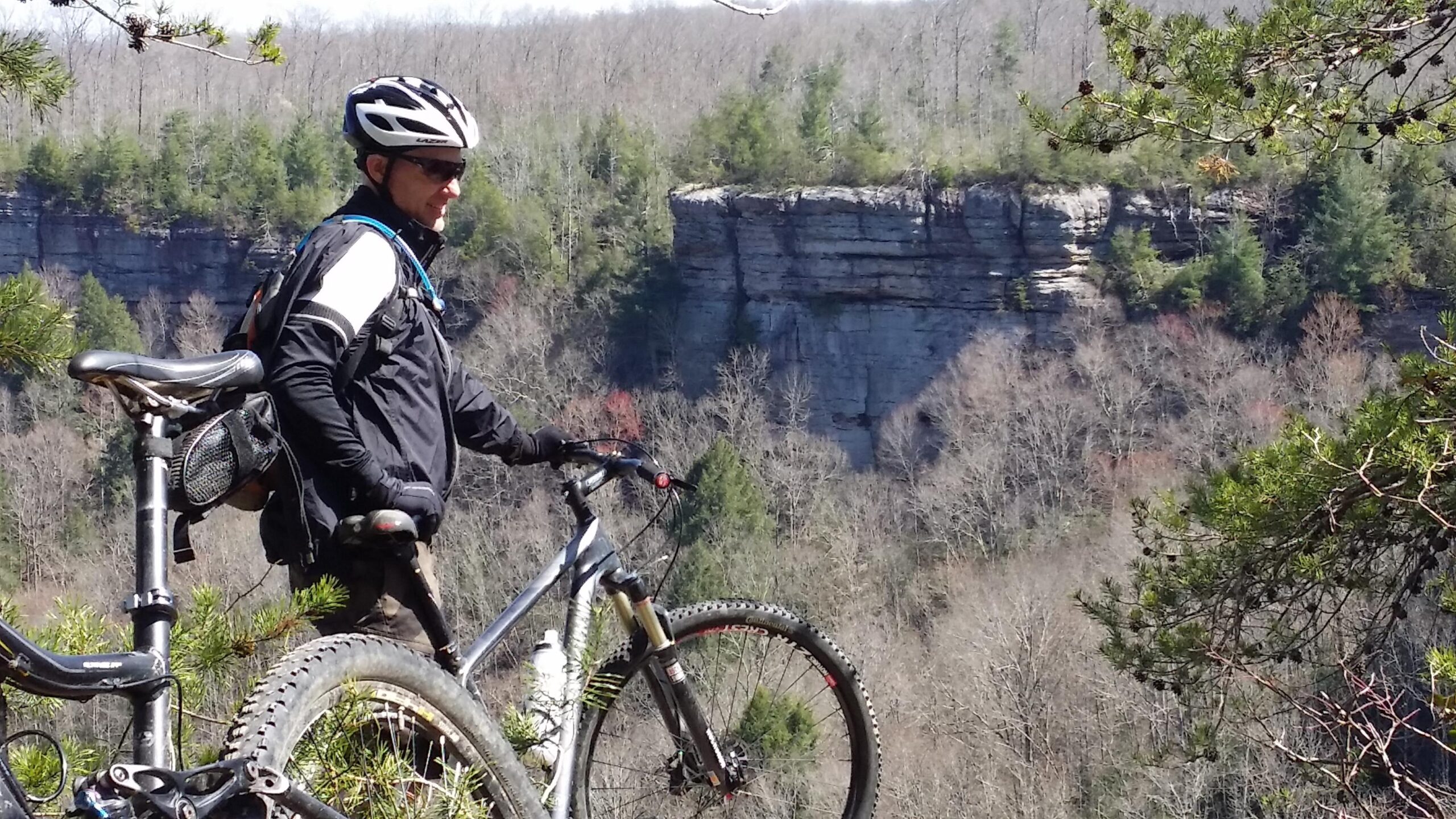 A mountain biker wearing a helmet and sunglasses stands next to his bike, gazing out over a scenic landscape featuring rocky cliffs and a forest of bare trees with some green conifers in the background. The scene captures the tranquility of nature during early spring. Big South Fork mountain bike trail.
