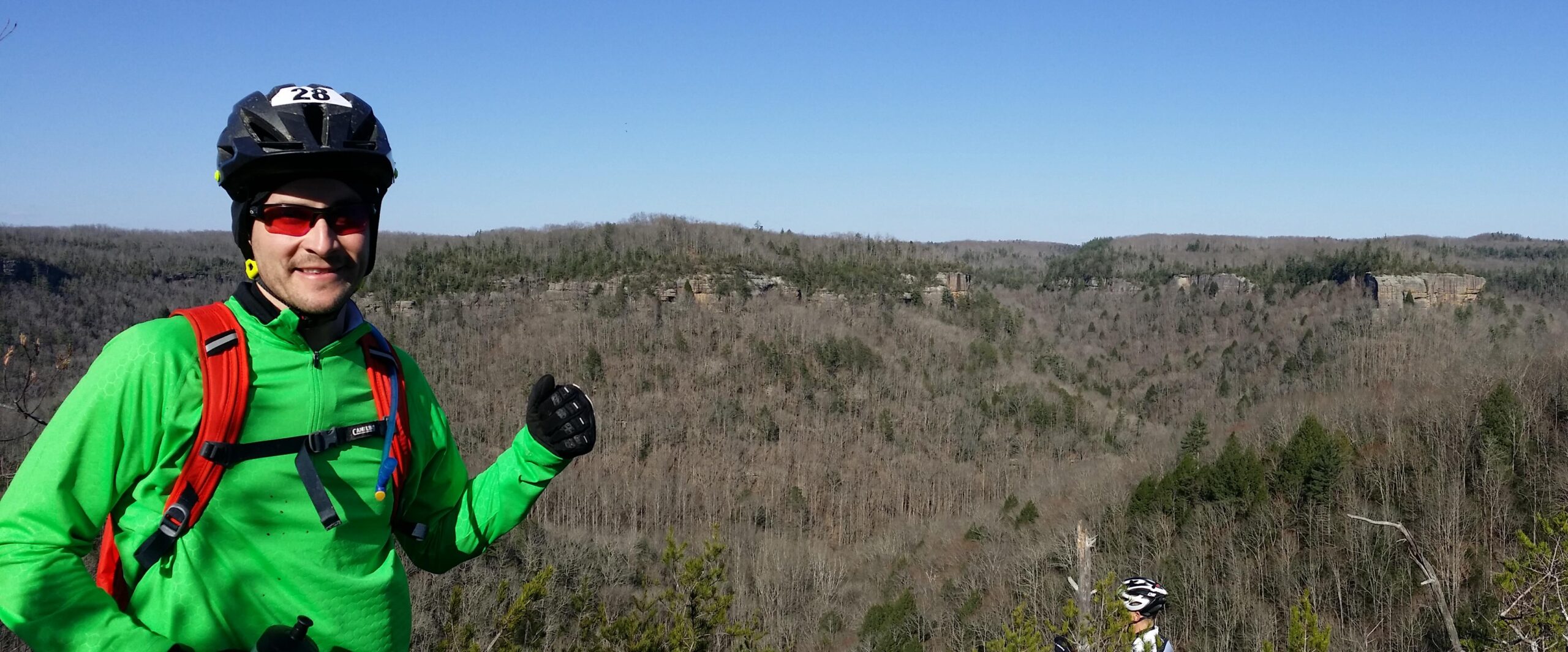 A man in a bright green cycling jacket and protective helmet stands on a viewpoint, smiling and gesturing with his right hand. Behind him, a panoramic view of a wooded valley can be seen, with rolling hills and cliffs under a clear blue sky. The image captures a moment of outdoor adventure and excitement. Big South Fork mountain bike trail.