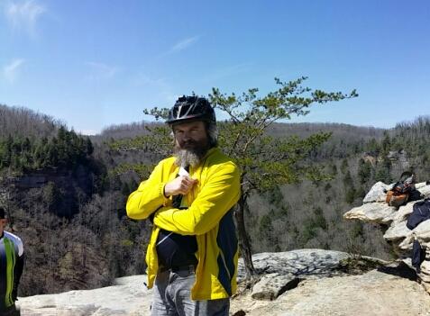A person wearing a yellow cycling jacket and a helmet stands confidently at the edge of a rocky cliff, surrounded by a forested landscape under a clear blue sky. Big South Fork mountain bike trail.