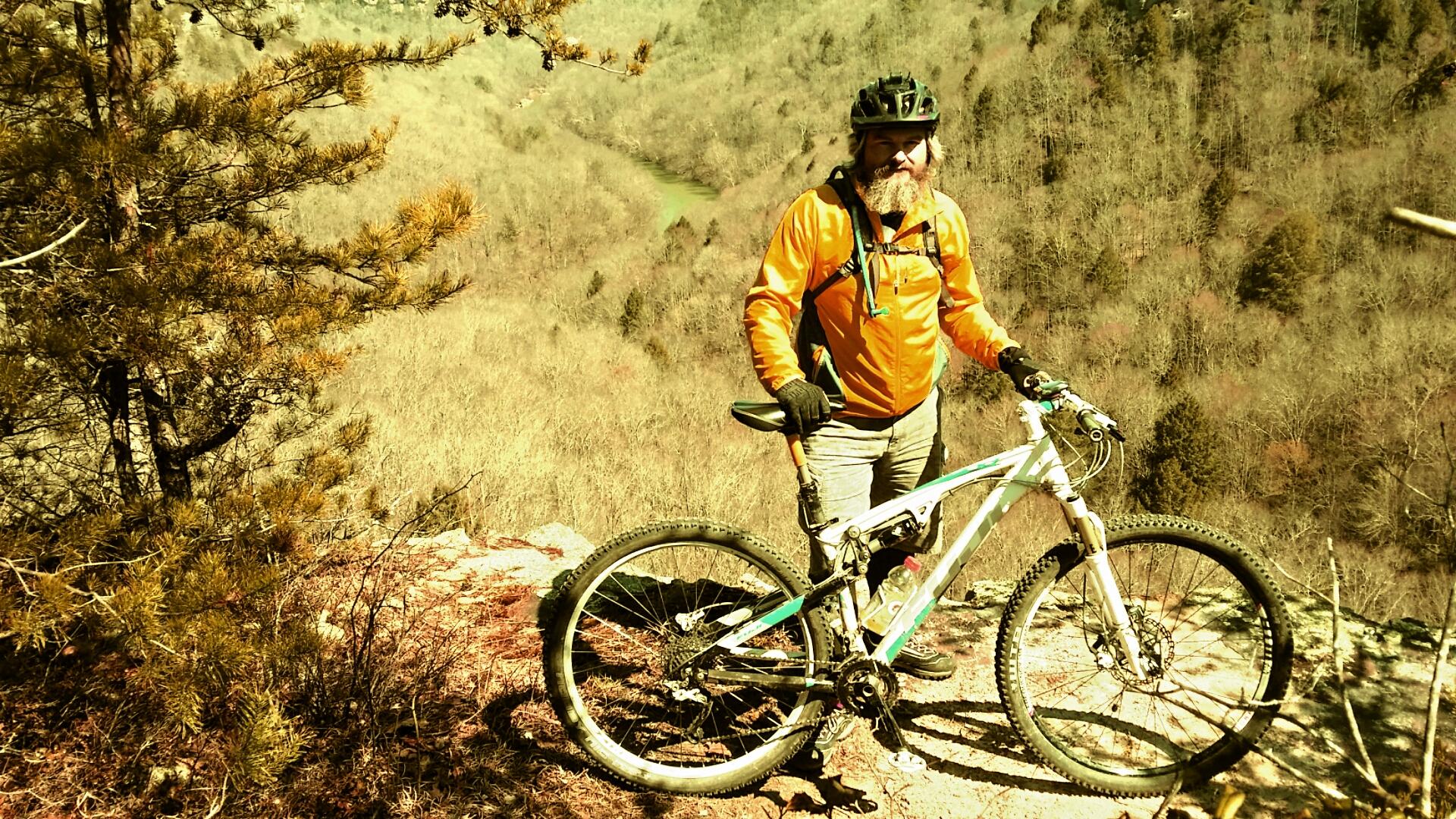 A mountain biker in an orange jacket and black gloves stands next to a white and teal mountain bike on a rocky ledge. The background features a scenic view of a valley with bare trees and rugged terrain under a clear sky. Big South Fork mountain bike trail.