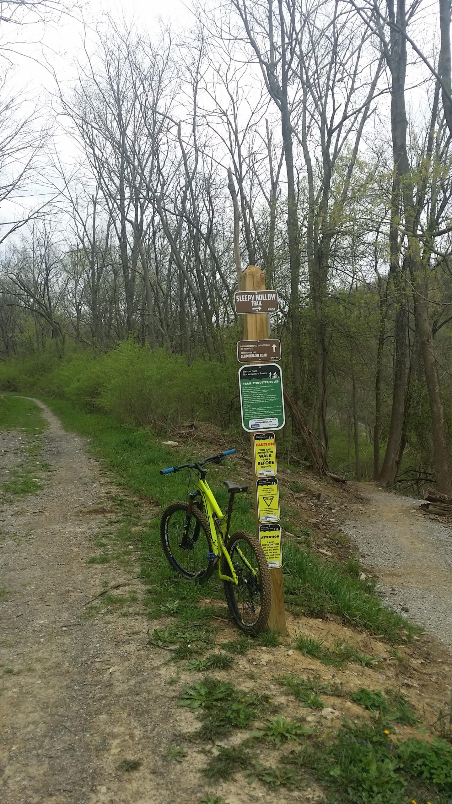 A vibrant yellow mountain bike is leaning against a wooden trail sign that reads "Sleepy Hollow Trail." The sign is accompanied by additional informational and safety notices. The setting features a gravel pathway winding through a wooded area with leafless trees and emerging greenery, indicating early spring. Devou Park mountain bike trail.