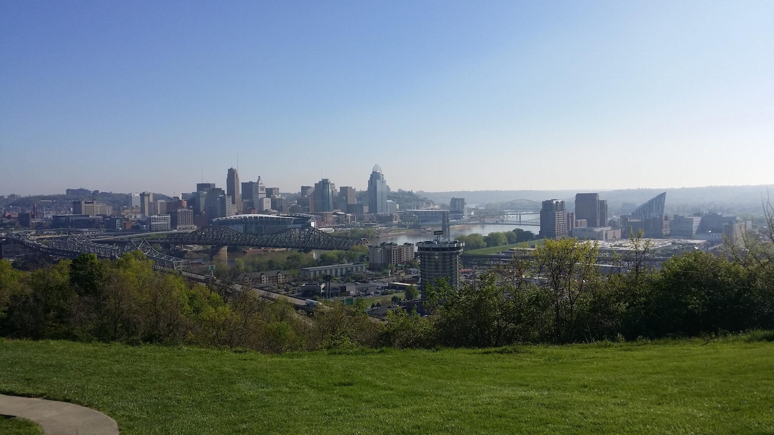 A panoramic view of a city skyline featuring various modern buildings, a bridge spanning a river, and lush greenery in the foreground under a clear blue sky. Devou Park mountain bike trail.