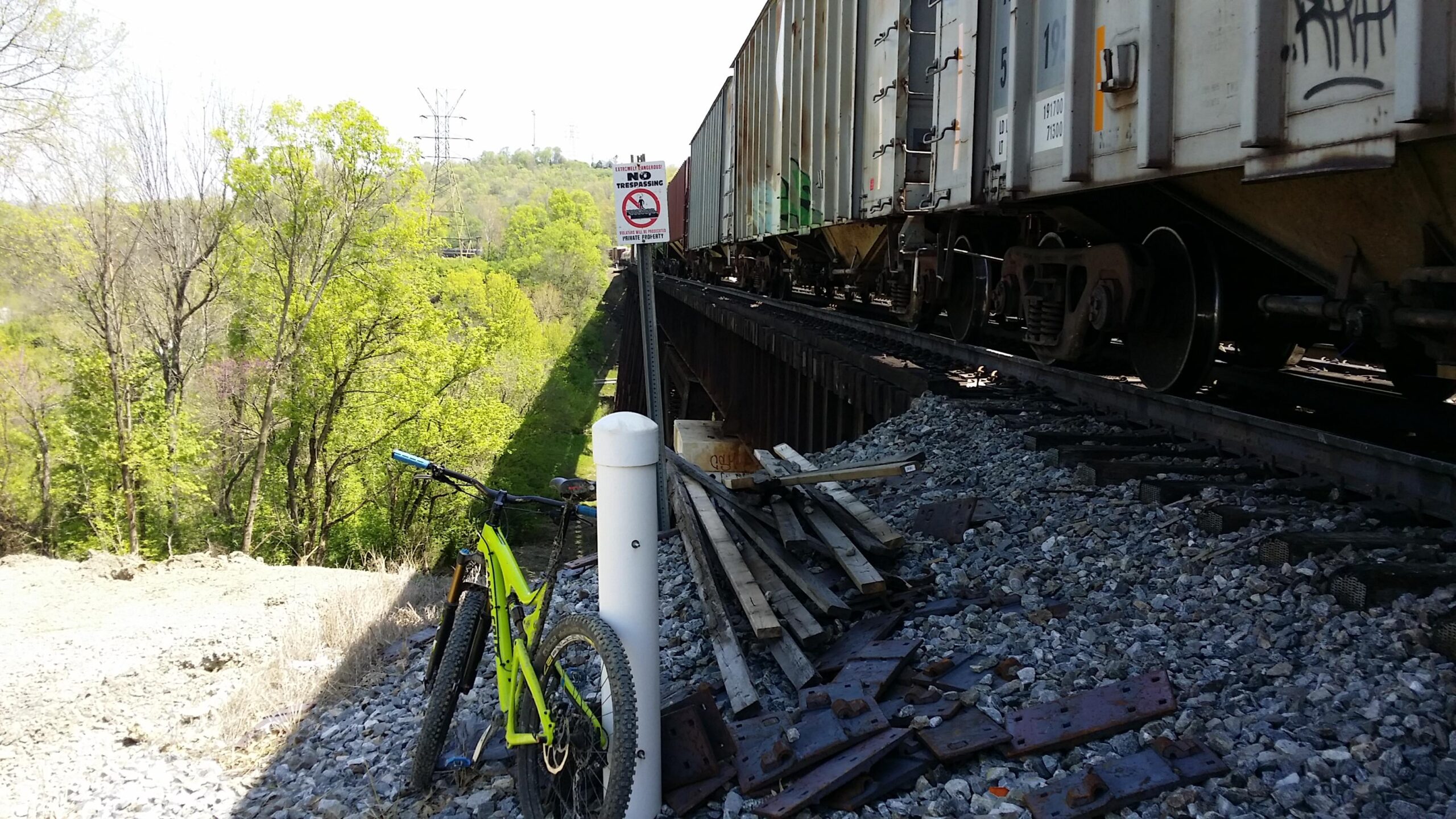 A bright green mountain bike rests on gravel beside a railroad track, with a freight train passing overhead. A "No Trespassing" sign is visible, and trees with fresh green leaves surround the area, indicating a spring setting. Rusted metal debris and wooden planks are scattered nearby. Devou Park mountain bike trail.