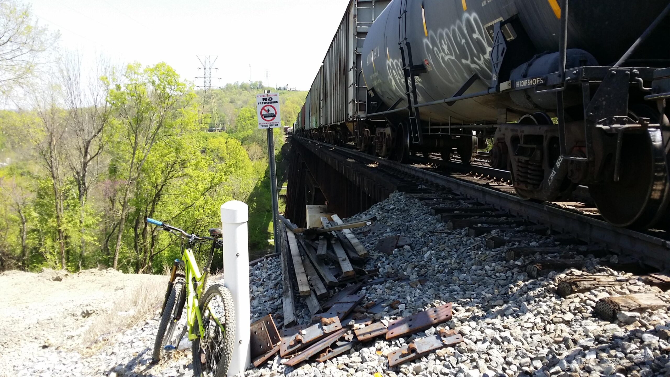 A green mountain bike rests against a white post near a railway track, surrounded by gravel and wooden debris. In the background, a freight train passes on an elevated track, with trees and power lines visible in the distance. A sign indicates "No Trespassing." Devou Park mountain bike trail.