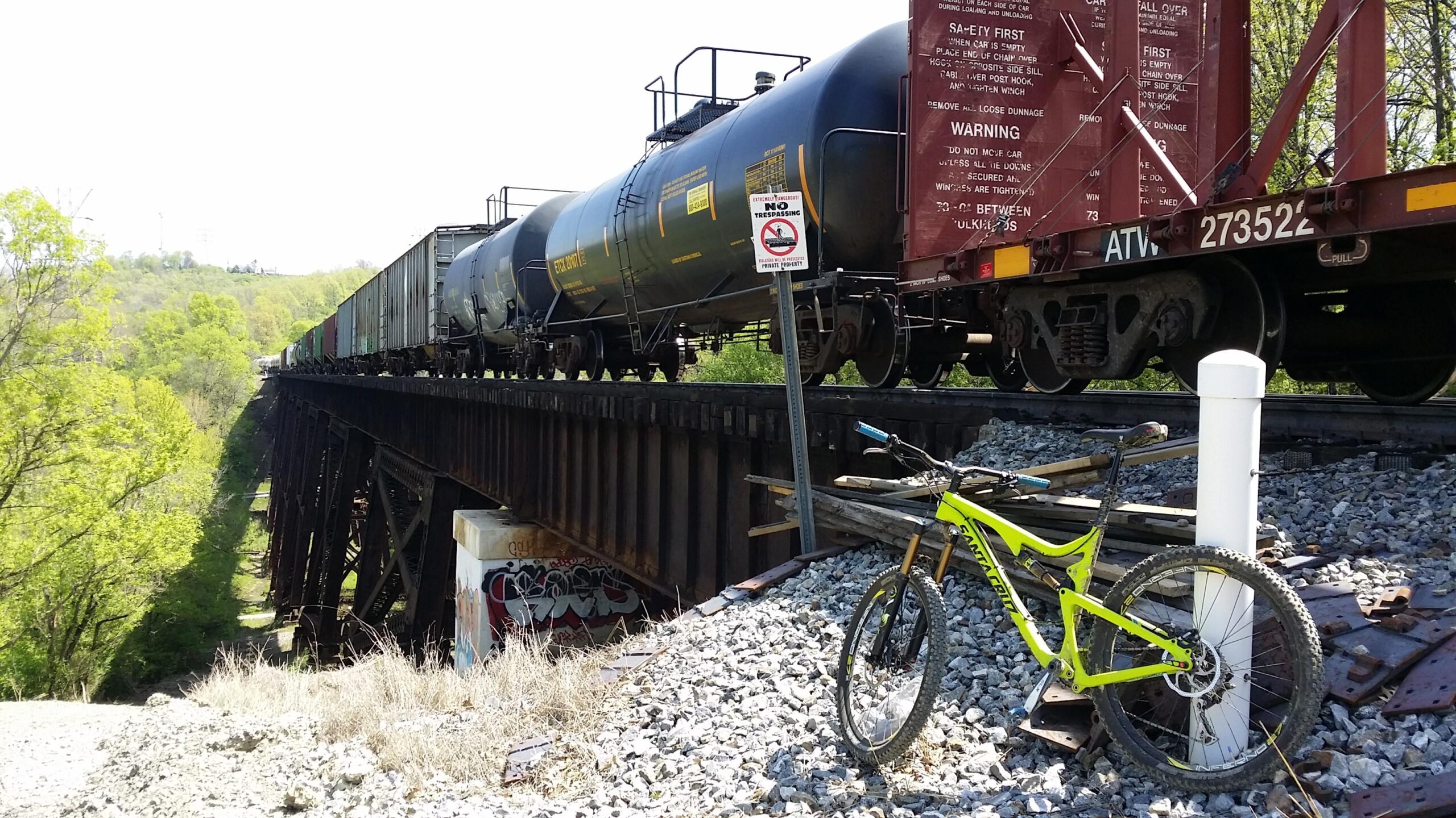 A mountain bike leans against a white post on rocky terrain next to a railway track. A freight train, composed of several cargo cars including a black tank car, is visible on an elevated trestle bridge surrounded by green foliage. A "No Trespassing" sign stands nearby, and graffiti is visible on one of the concrete supports. Devou Park mountain bike trail.