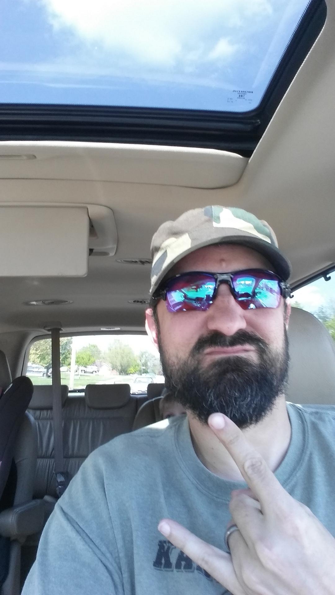 A person sitting in the driver's seat of a vehicle, wearing a camouflage cap and sunglasses, making a hand gesture. The interior of the car is visible, as well as a glimpse of a child in a car seat in the background. The sky is visible through the sunroof above. Devou Park mountain bike trail.