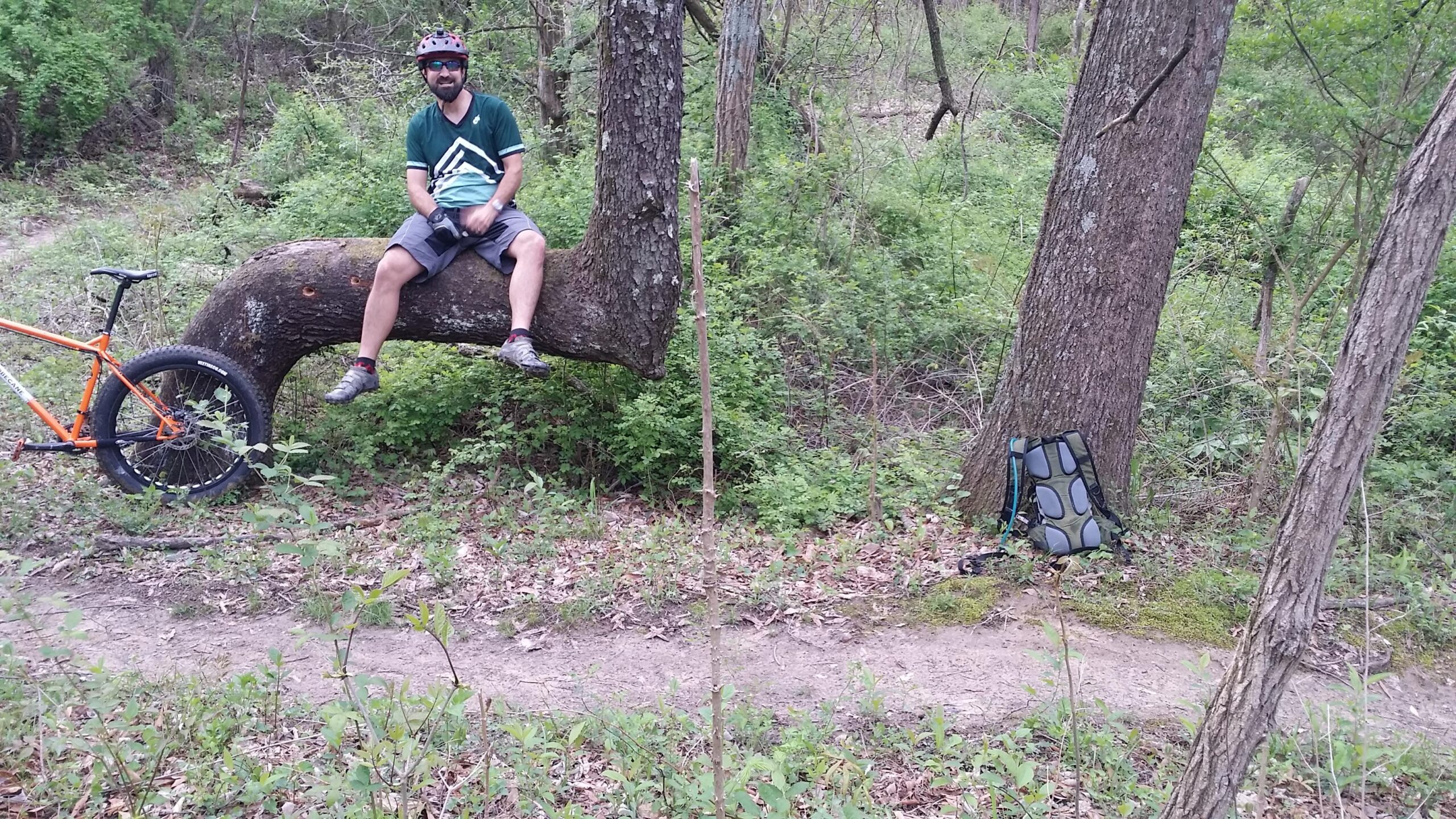 A man wearing a helmet and cycling gear sits on a large, horizontal tree branch in a lush green forest. An orange mountain bike is propped against the tree, and a backpack is resting on the ground nearby. A dirt trail is visible in the background, surrounded by greenery and undergrowth. Versailles State Park mountain bike trail.