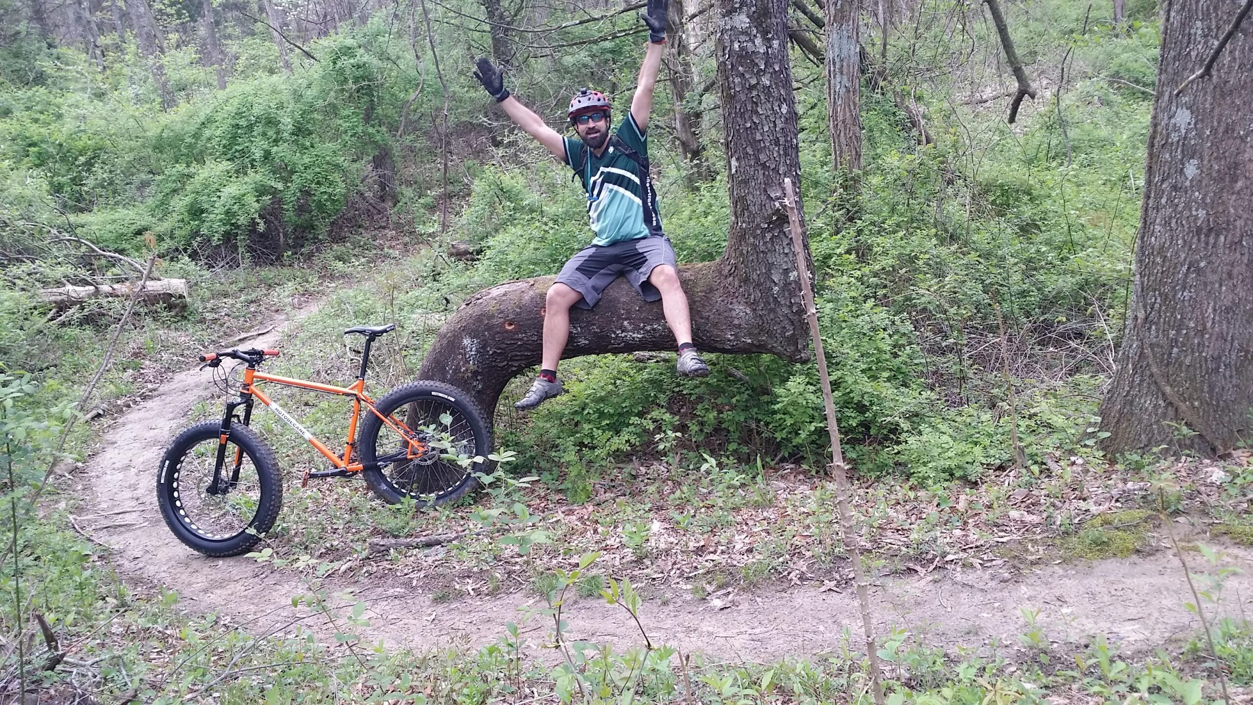 A person sitting on a curved tree branch in a forested area, with arms raised in a joyful pose. An orange mountain bike is parked on a dirt trail nearby, surrounded by green foliage and trees. Versailles State Park mountain bike trail.