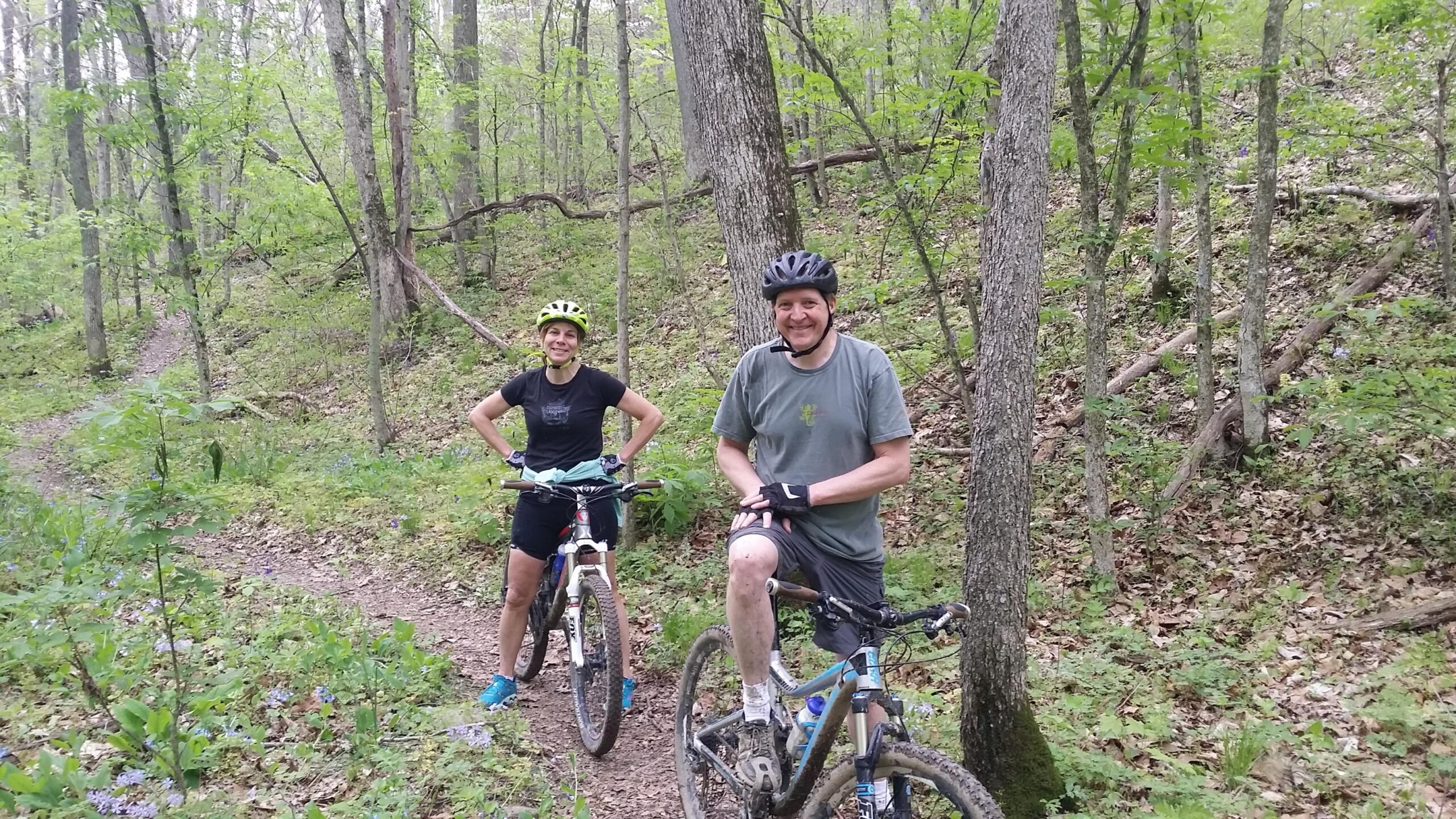 Two mountain bikers pause on a trail in a lush, green forest. One rider is standing beside their bike, smiling, while the other is sitting on their bike, wearing a helmet and gloves. The scene features a natural setting with trees, greenery, and a winding dirt path in the background. Versailles State Park mountain bike trail.