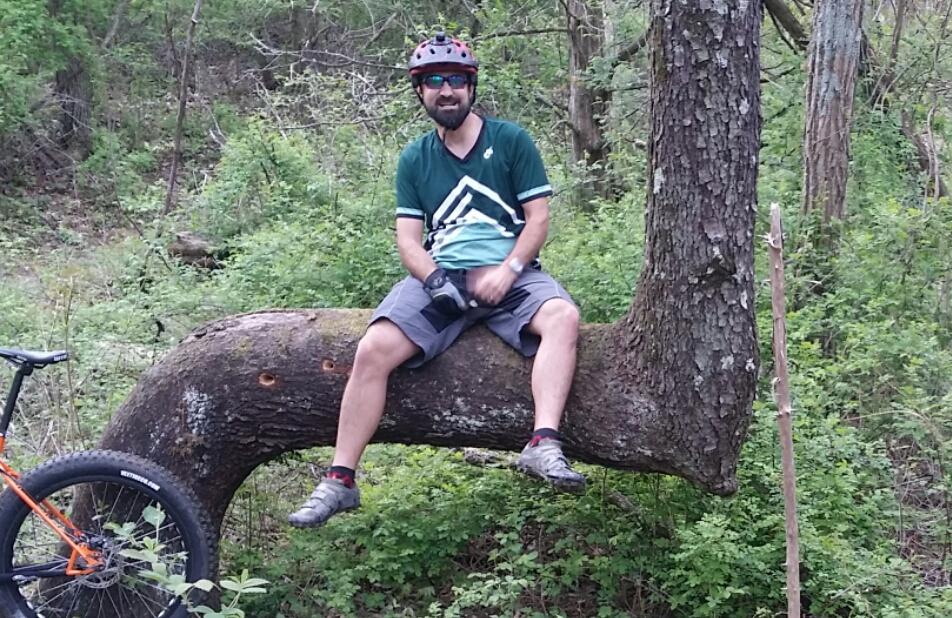 A person sitting on a large, bent tree trunk in a green, wooded area, wearing a bicycle helmet and a teal and white athletic shirt. A mountain bike is leaning against the tree nearby. Versailles State Park mountain bike trail.