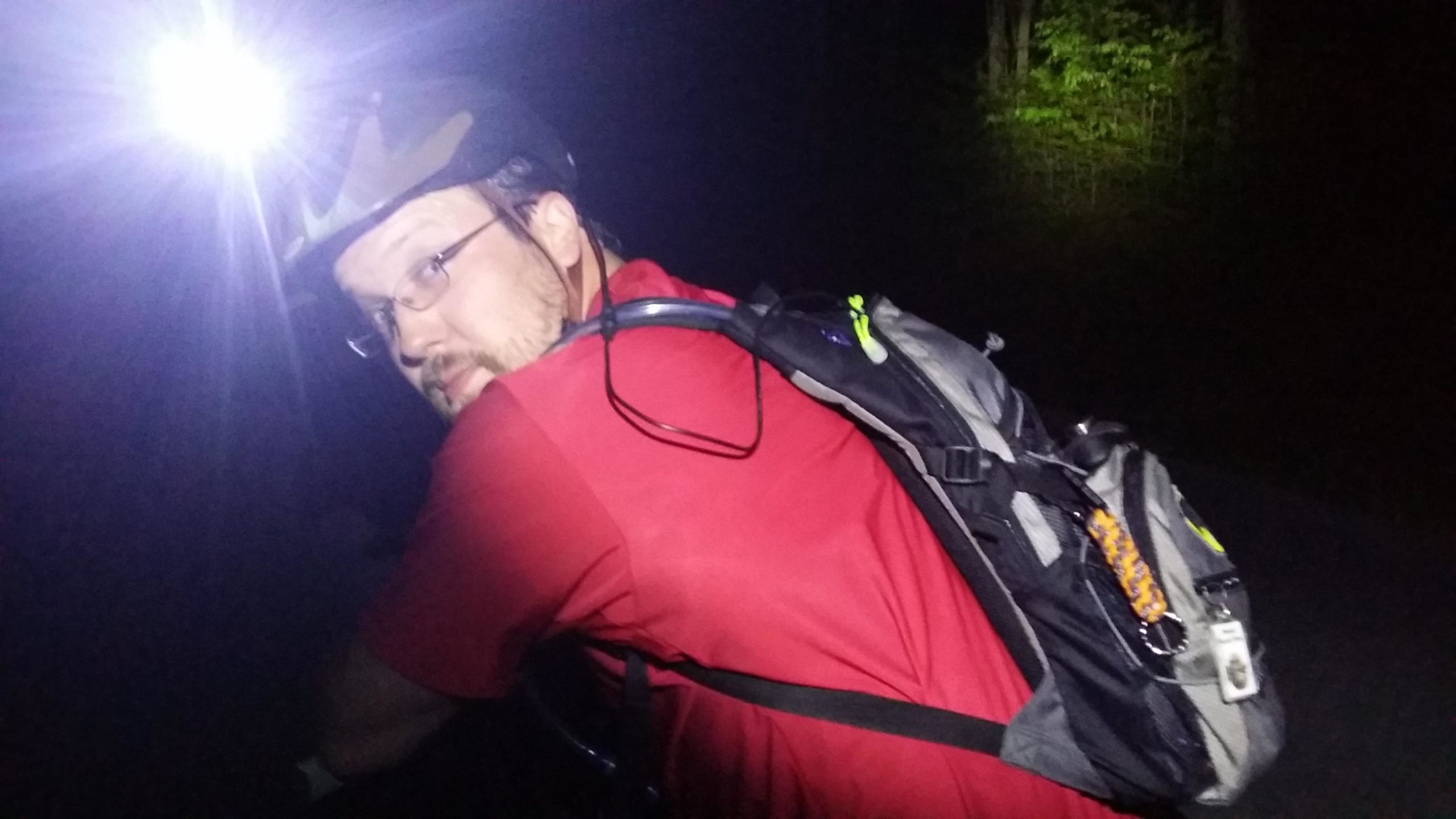 A person wearing a helmet and a red shirt, with a backpack, is riding a bicycle at night. A bright headlight illuminates the scene, casting light on their face and surroundings. The background is dark, with hints of greenery visible in the distance. Sheltowee Trace - Laurel Lake Trail mountain bike trail.