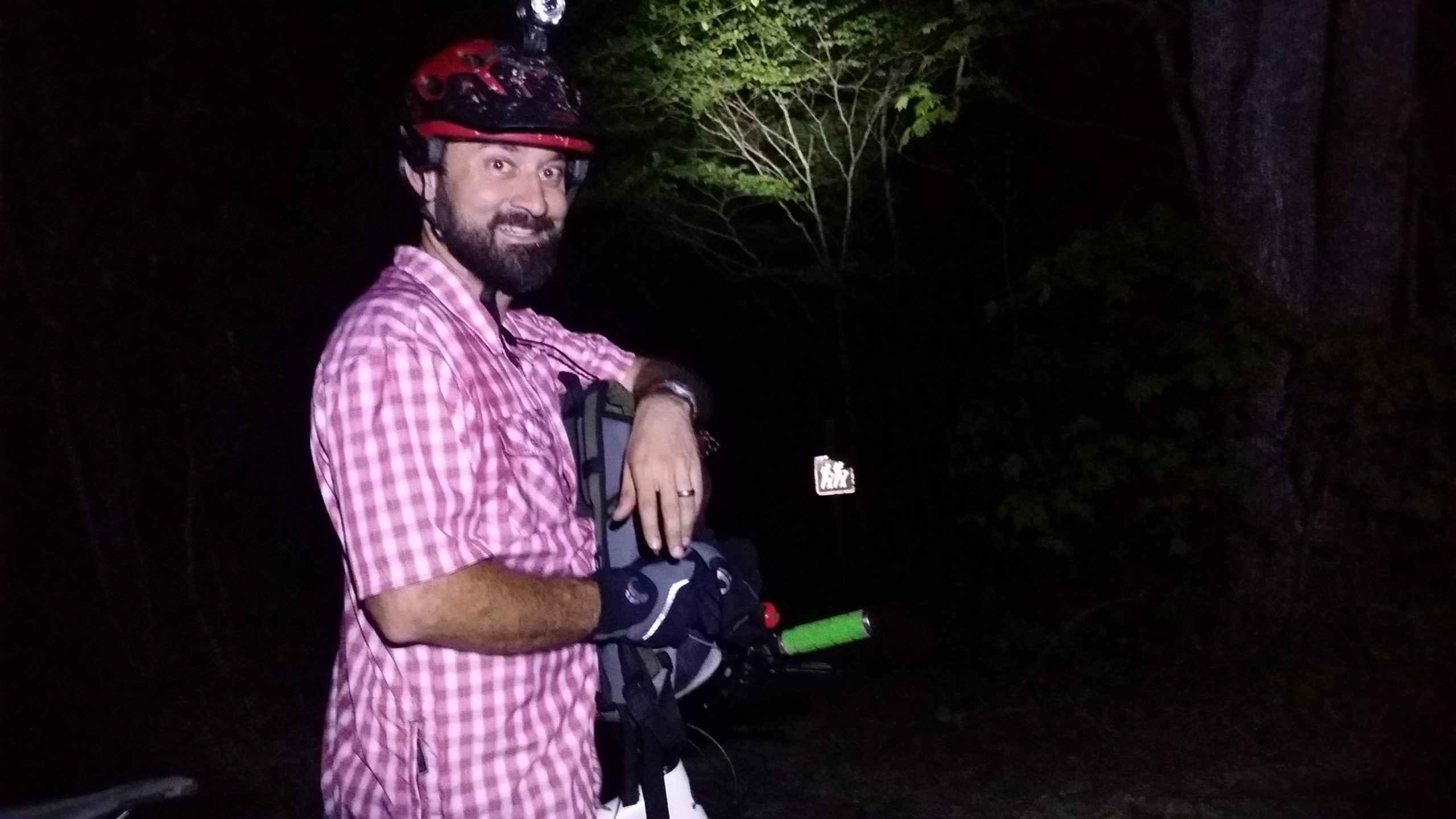 A person wearing a red helmet with a headlamp stands in a dimly lit area, smiling and leaning against a bicycle. They are dressed in a pink checkered shirt and black gloves, holding a flashlight with a green handle. The background features trees and a low-visibility sign in the distance. Sheltowee Trace - Laurel Lake Trail mountain bike trail.