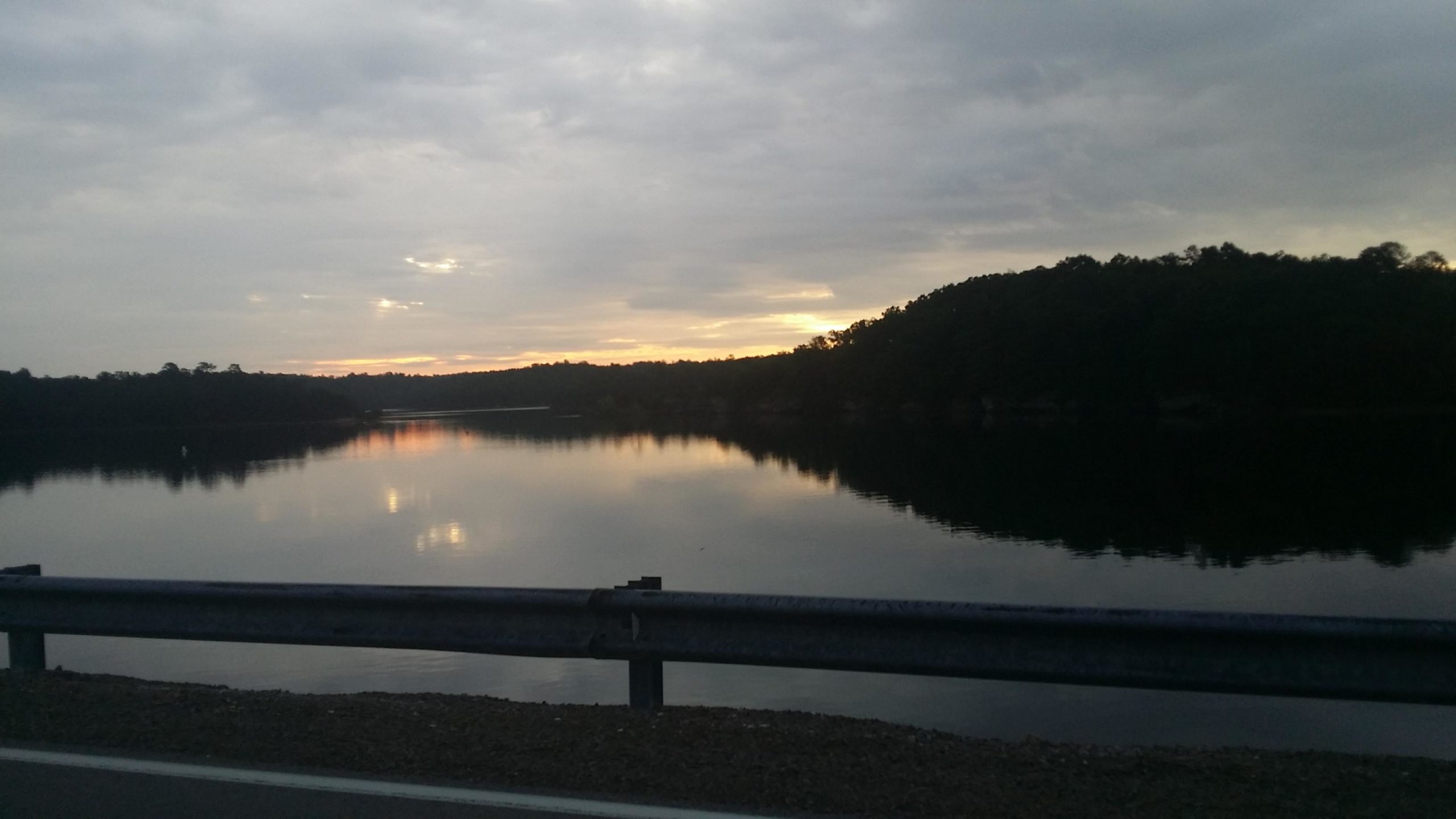 A tranquil lakeside scene at sunset, featuring calm water reflecting the cloudy sky and distant trees. A road barrier lines the foreground. Sheltowee Trace - Laurel Lake Trail mountain bike trail.