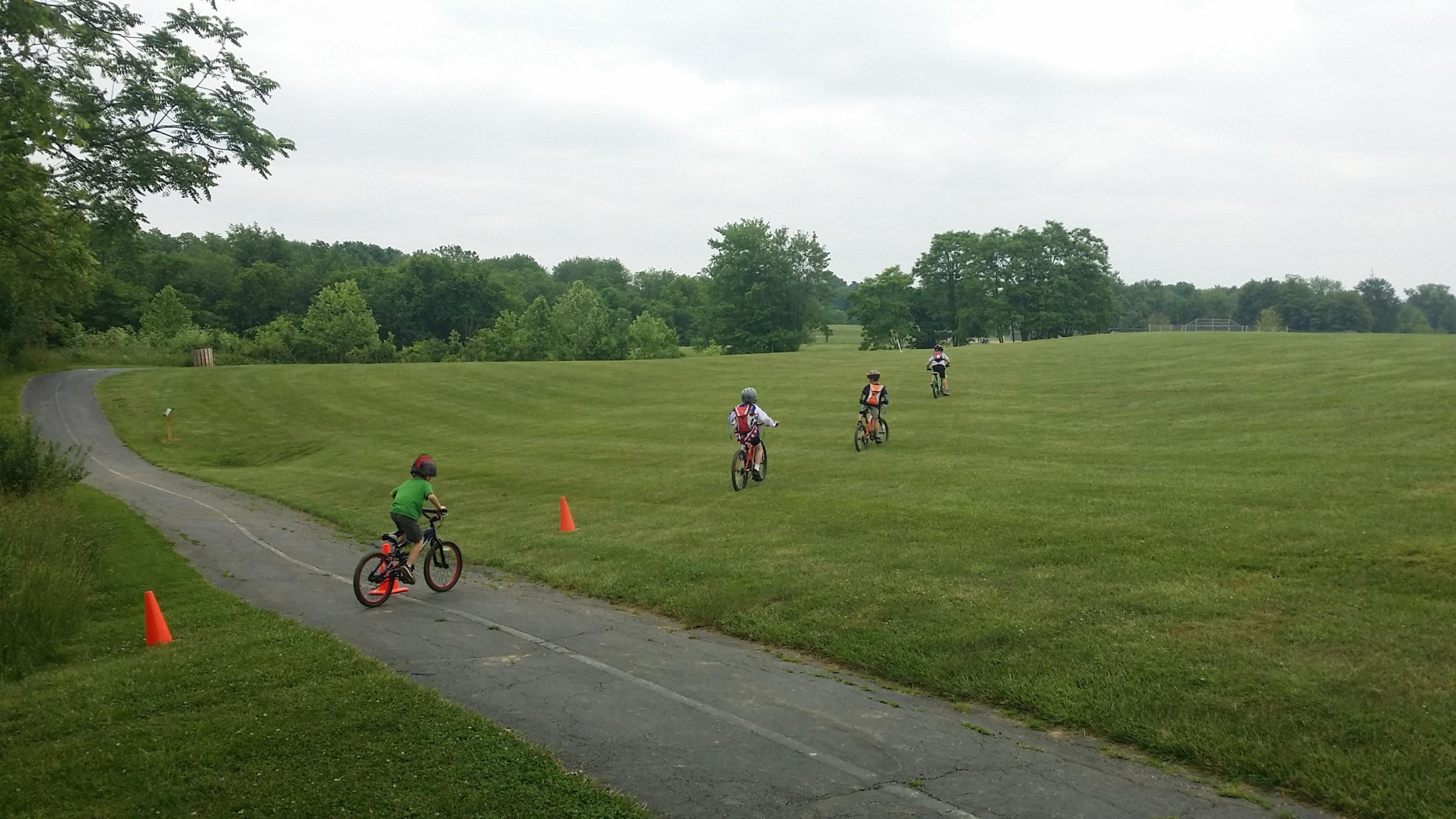 A group of children is riding bicycles on a paved path that winds through a grassy park. One child in a green shirt and helmet is maneuvering around a bright orange cone, while three other children are further ahead on the path, enjoying their ride. The park features lush green trees in the background, under a cloudy sky. England Idlewild Mountain Biking Park mountain bike trail.