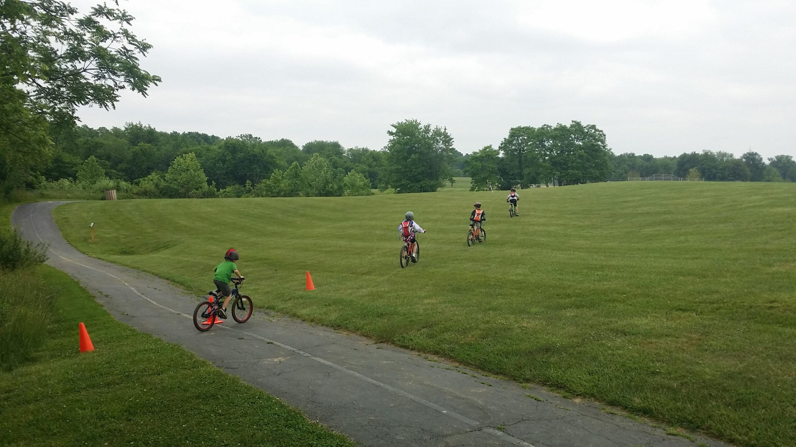Four children riding bicycles on a grassy field, with one child navigating a path near orange traffic cones. Green trees and a cloudy sky are visible in the background. England Idlewild Mountain Biking Park mountain bike trail.
