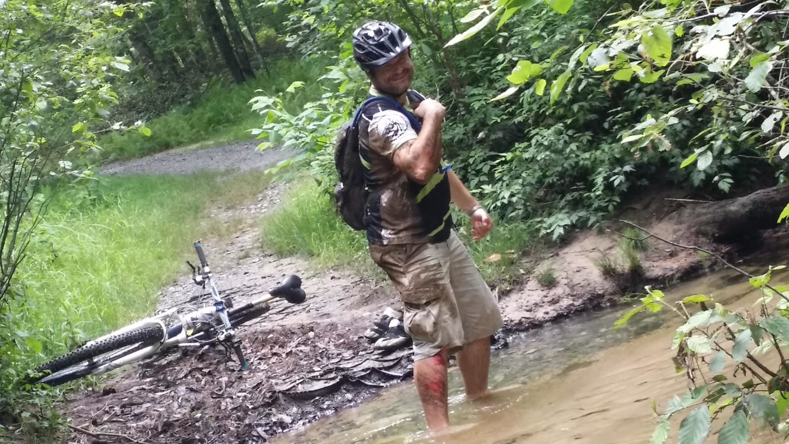 A person standing in shallow water along a wooded trail, smiling and wearing a helmet and a backpack. They are covered in dirt, indicating they have been biking or hiking in muddy conditions. A bicycle is lying on its side in the background, partially obscured by vegetation. DuPont State Recreational Forest mountain bike trail.