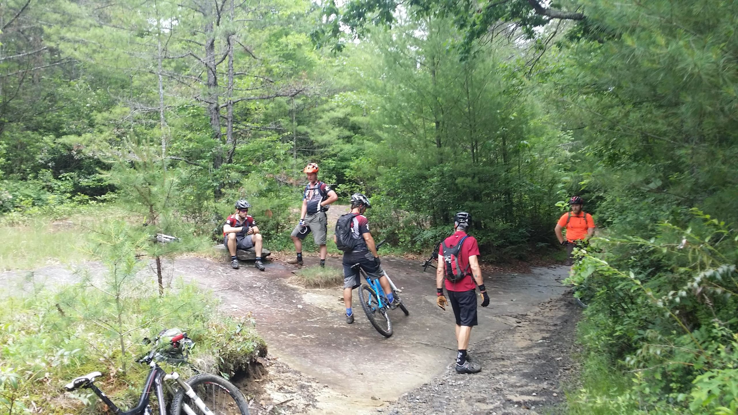 A group of six mountain bikers resting on a rocky trail surrounded by lush green trees. Some riders are seated, while others are standing with their bikes, engaged in conversation. The atmosphere is casual and relaxed, reflecting a break during their biking activity. DuPont State Forest mountain bike trail.