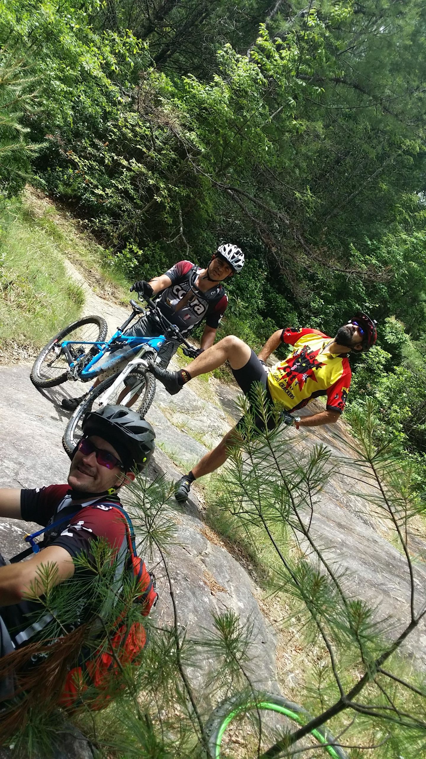 Three cyclists in mountain biking gear are gathered on a rocky trail surrounded by greenery. One cyclist sits on the ground, smiling, while another stands beside a blue mountain bike with one leg raised. The third cyclist, wearing a colorful jersey, strikes a playful pose with hands on hips. Sunlight filters through the trees, highlighting the outdoor setting. DuPont State Recreational Forest mountain bike trail.