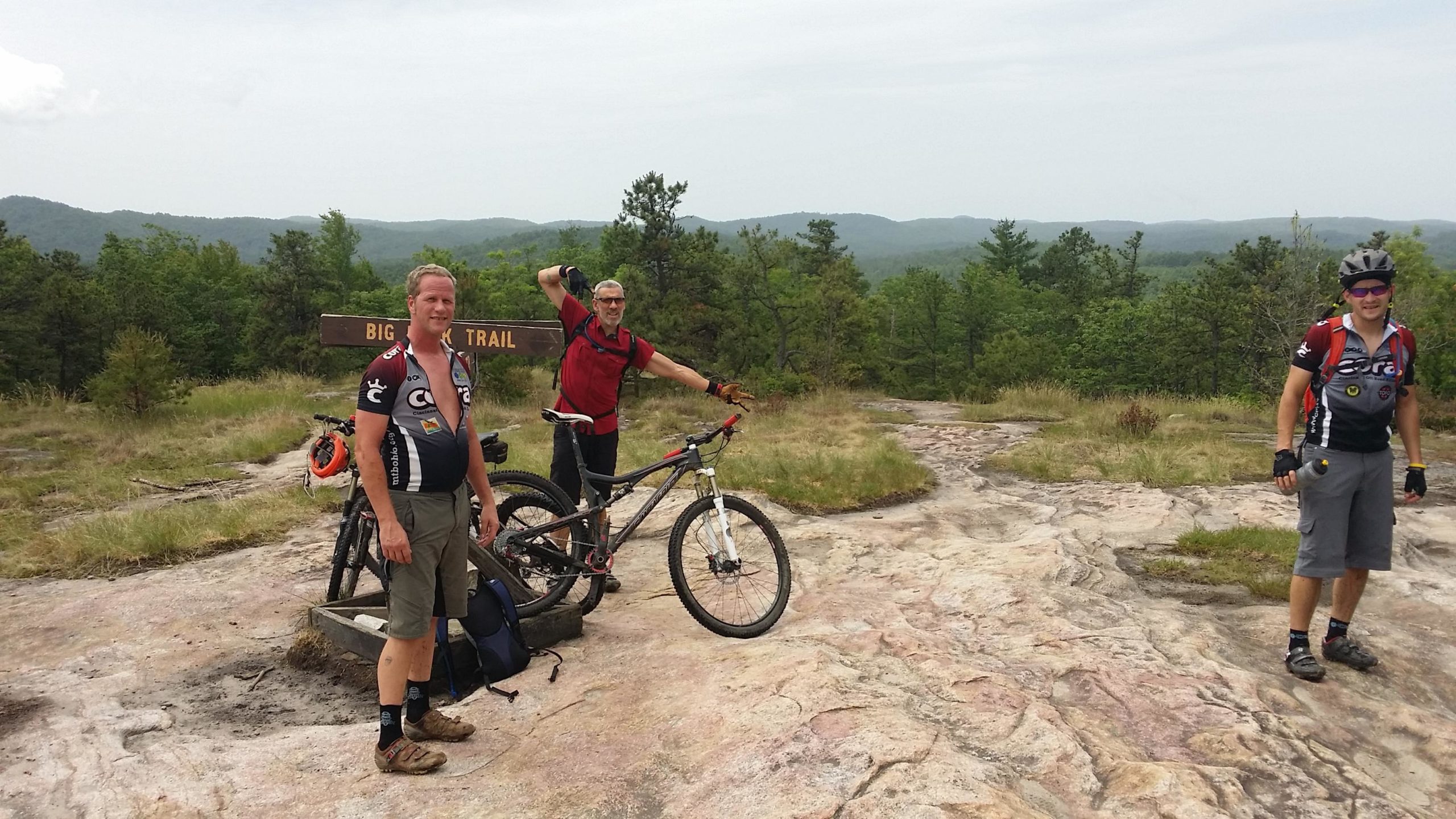 Three mountain bikers stand at the Big Rock Trail sign, surrounded by a scenic landscape of rolling hills and greenery. One cyclist is posing enthusiastically with his arm raised, while the others stand beside their bikes, wearing cycling gear. The rocky terrain and natural backdrop create an outdoor recreational atmosphere. DuPont State Recreational Forest mountain bike trail.