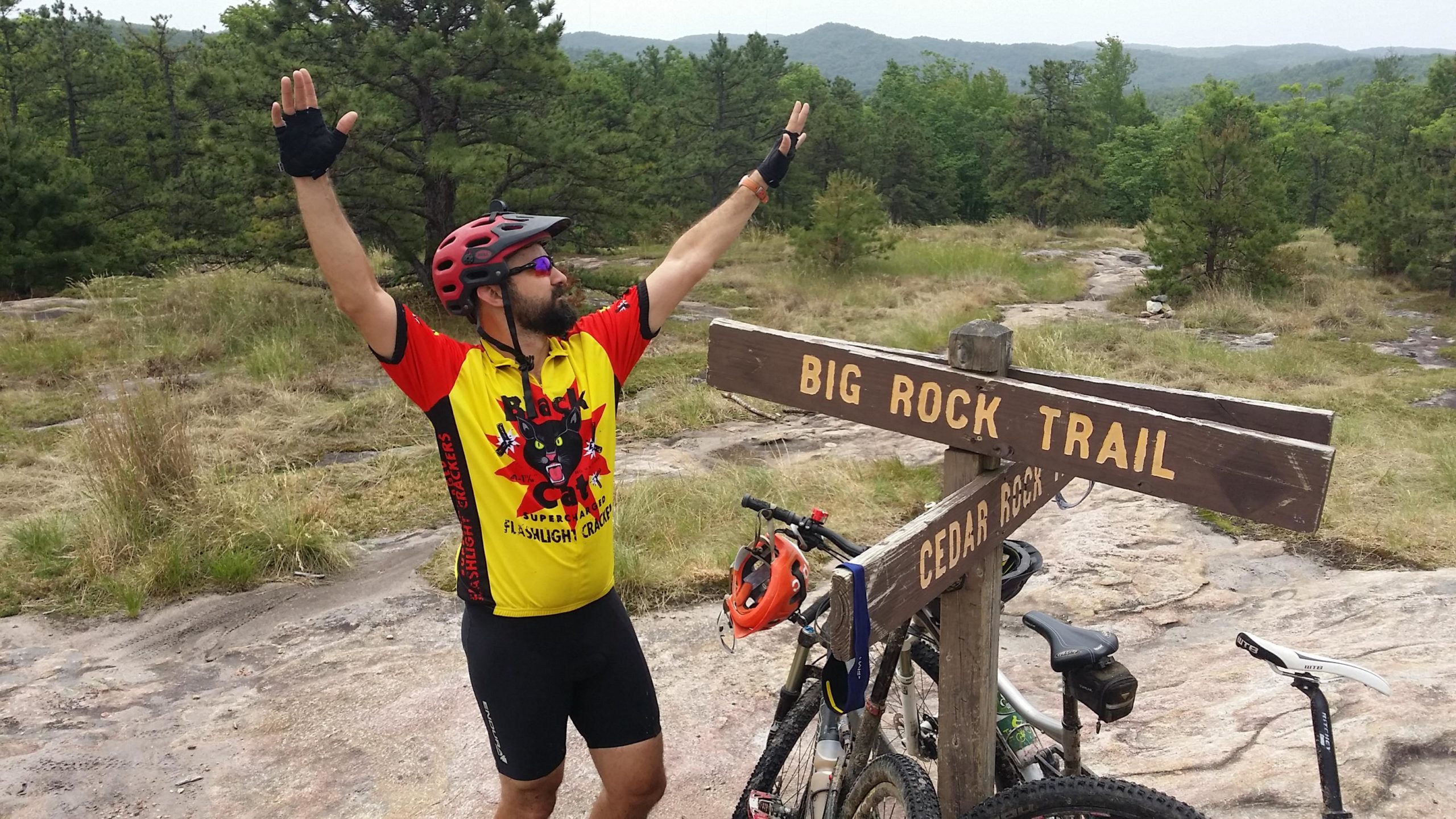 A mountain biker in a colorful jersey stands beside a wooden sign marked "Big Rock Trail," raising one arm in triumph. The backdrop features a lush, green landscape with trees and hills. Two bicycles are positioned next to the sign on rocky terrain, suggesting a challenging trail experience. DuPont State Forest mountain bike trail.