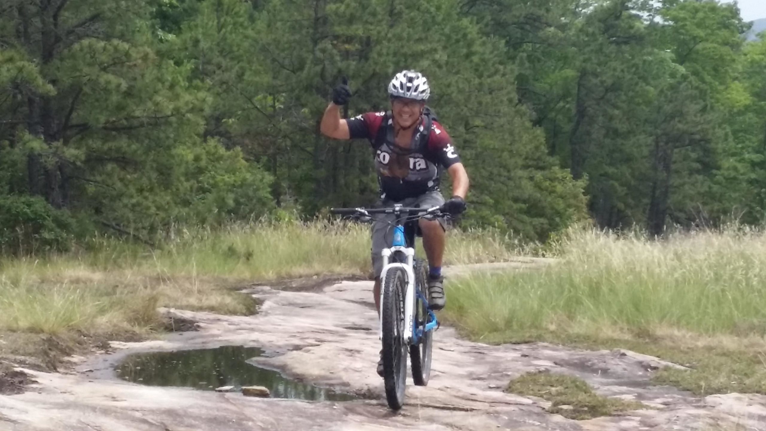 A person riding a mountain bike on a rocky trail, smiling and giving a thumbs-up. Lush greenery and trees are visible in the background, with patches of grass and a small puddle near the path. DuPont State Recreational Forest mountain bike trail.