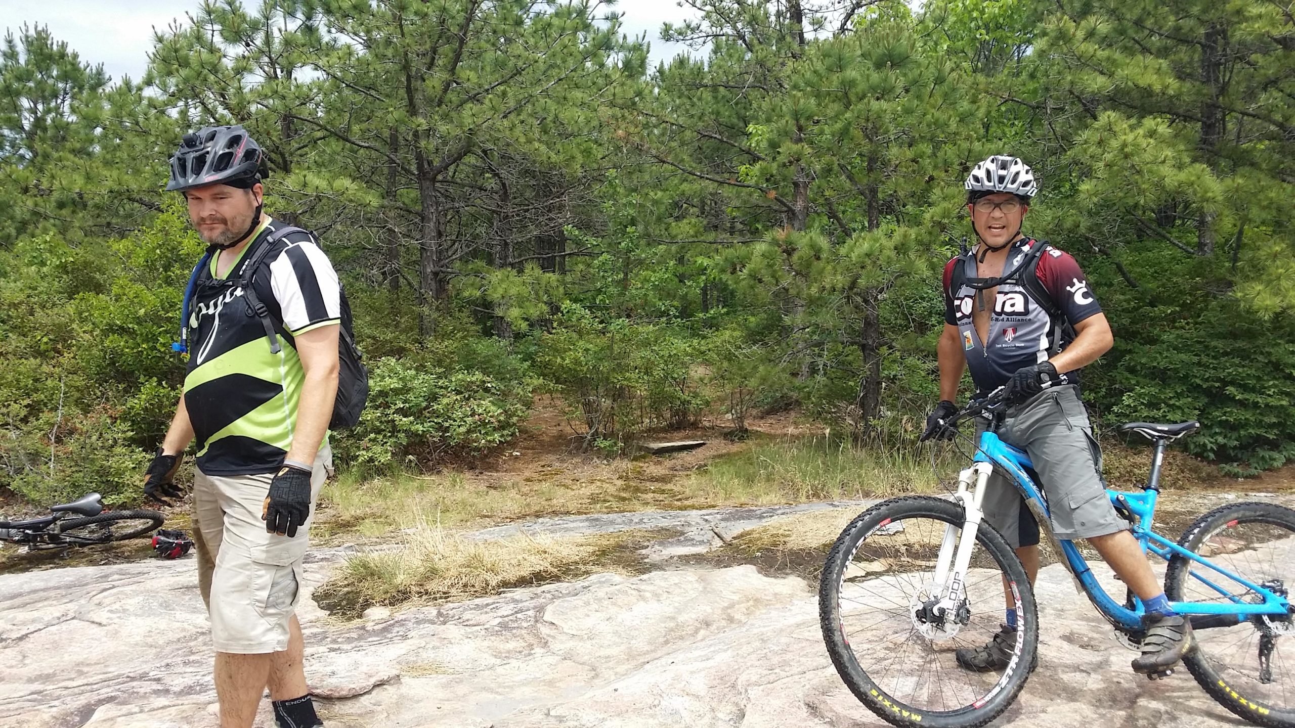Two mountain bikers are standing on a rocky outdoor trail surrounded by trees. One is wearing a black and green jersey with gloves and a helmet, while the other is in a dark jersey with a backpack, also wearing gloves and a helmet. They appear to be taking a break, with bikes parked nearby and a natural landscape in the background. DuPont State Forest mountain bike trail.