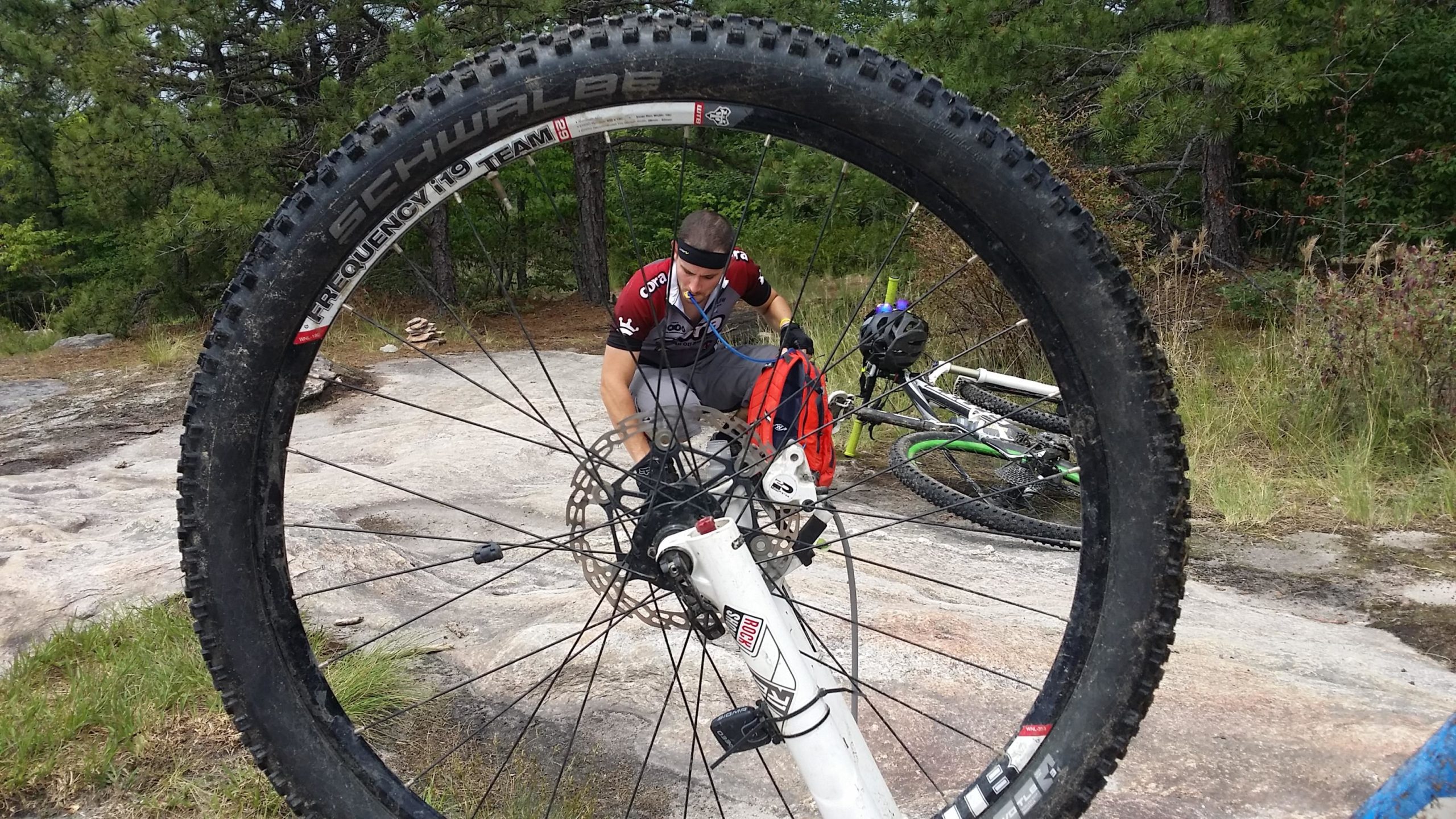 A mountain biker kneeling down to inspect a bicycle wheel, with the tire in the foreground. The setting features rocky terrain and greenery in the background, showcasing an outdoor biking environment. DuPont State Forest mountain bike trail.