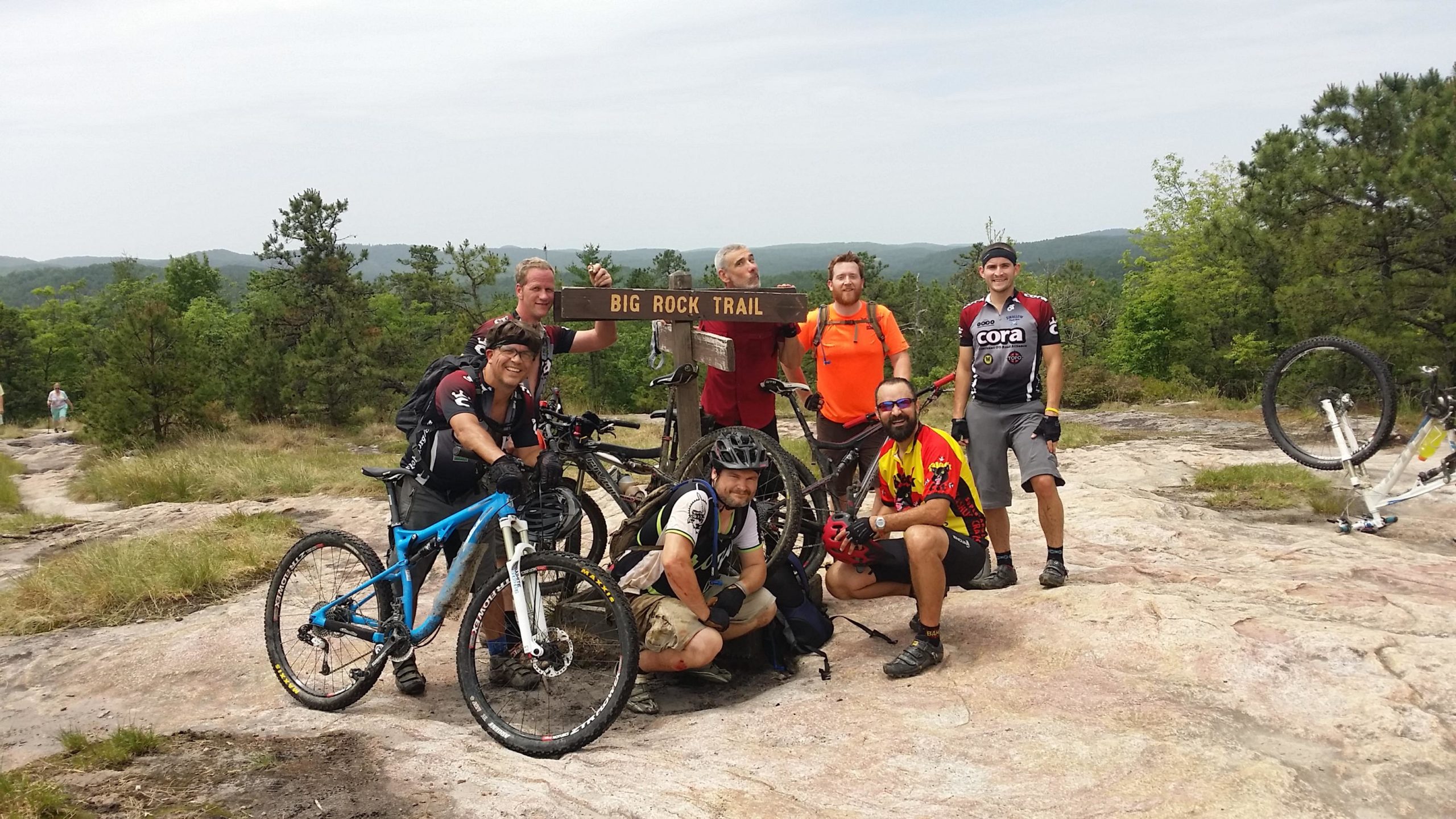 A group of seven mountain bikers posing at the Big Rock Trail sign in a scenic outdoor setting, surrounded by greenery and hills. They are wearing cycling gear and helmets, with their bicycles nearby. The atmosphere is cheerful and adventurous. DuPont State Forest mountain bike trail.