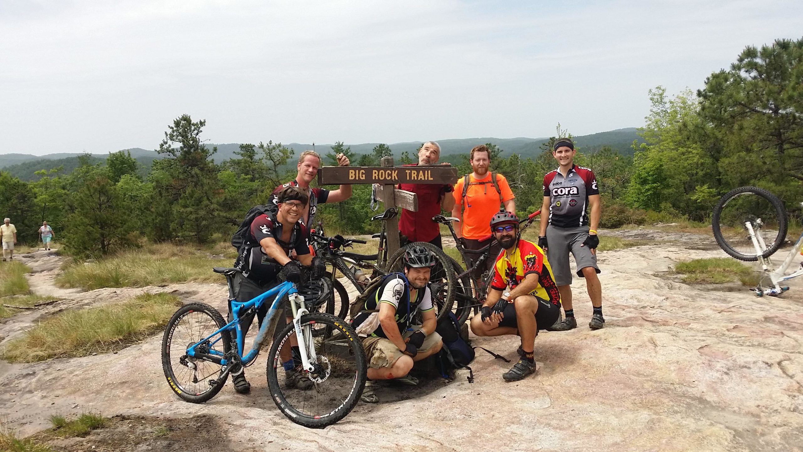 A group of seven mountain bikers posing in front of a wooden sign that reads "Big Rock Trail." The background features a greenery-filled landscape with hills, while two hikers can be seen on a path in the distance. The bikers are dressed in cycling gear and helmets, with several bikes resting nearby. DuPont State Forest mountain bike trail.