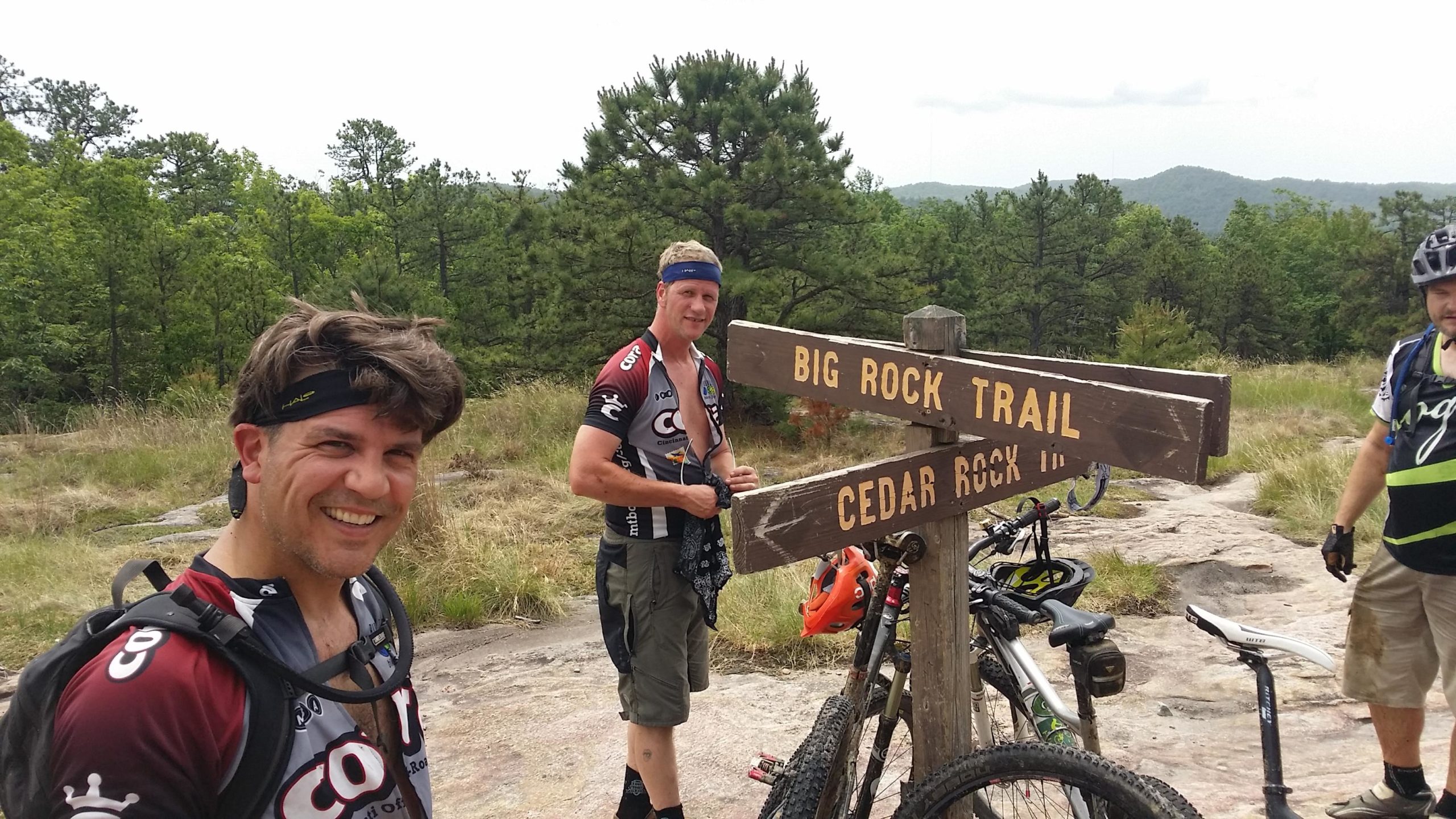 A group of three mountain bikers posing near a wooden trail sign that reads "Big Rock Trail" and "Cedar Rock IV." They are on a rocky path surrounded by greenery, with mountain bikes leaning against the sign. The riders are dressed in cycling gear, enjoying a break during their ride. DuPont State Forest mountain bike trail.