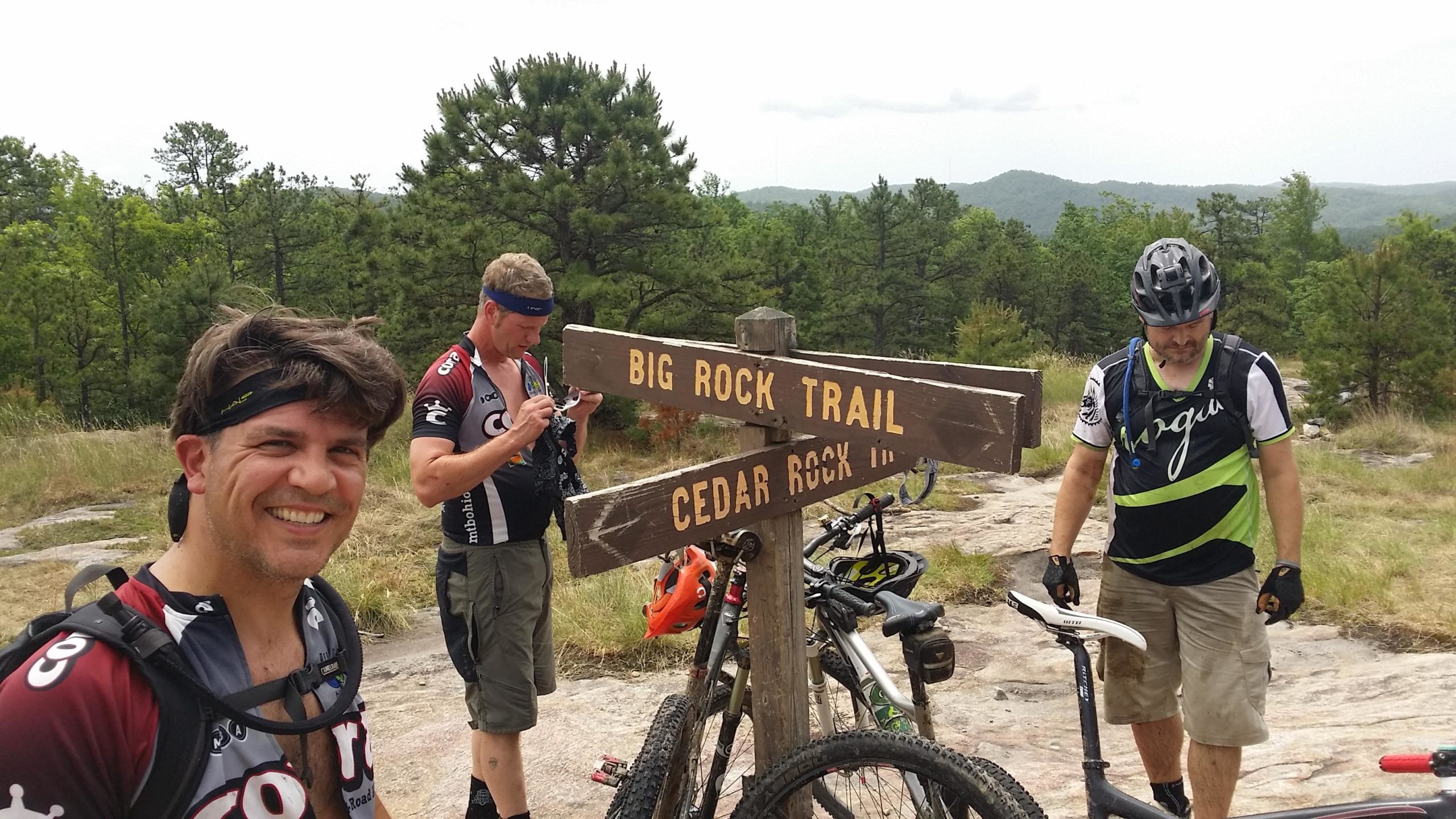 Three mountain bikers pause at a trail sign indicating directions to "Big Rock Trail" and "Cedar Rock In." In the background, lush greenery and hills are visible under a cloudy sky. One biker is smiling at the camera, while the others check their gear, surrounded by parked bicycles. DuPont State Recreational Forest mountain bike trail.