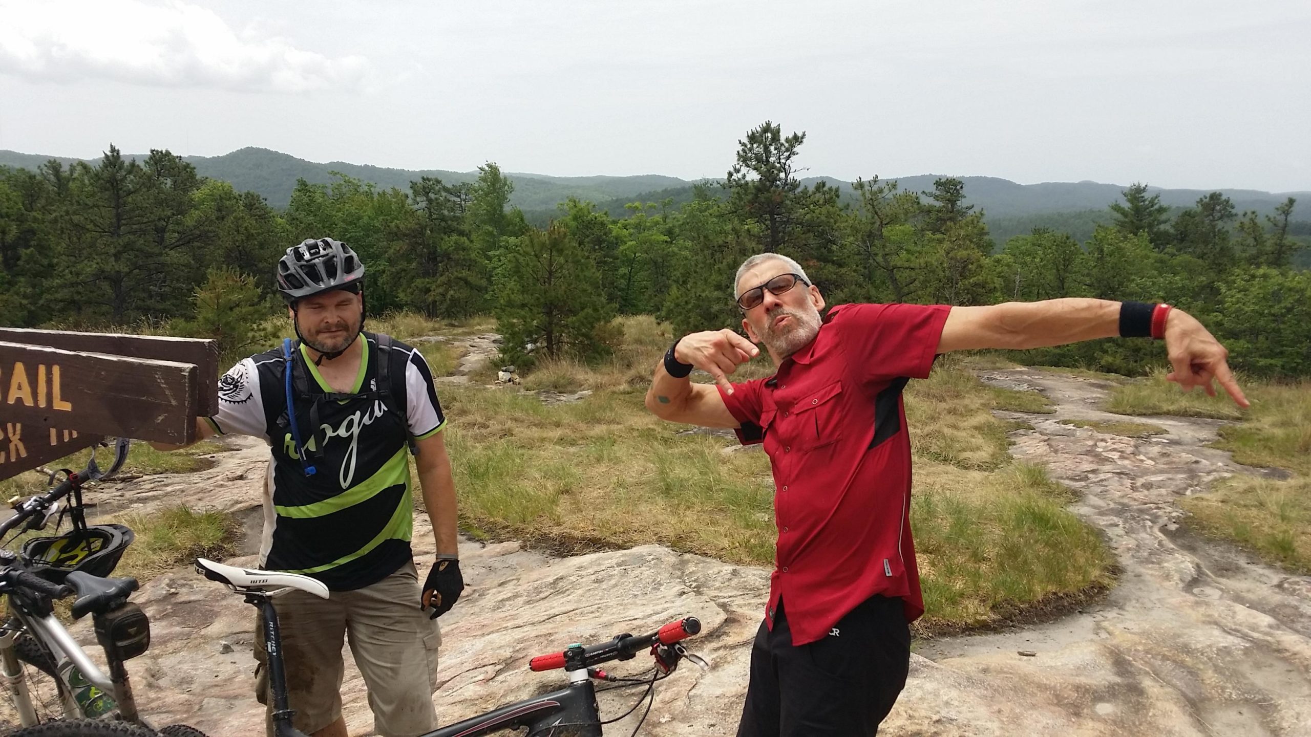 Two men are standing on a rocky outcrop with a lush green landscape and distant hills in the background. One man, in a black and green biking jersey, is next to a mountain bike, holding a sign that reads "TRAIL." The other man, wearing a red shirt and glasses, is posing playfully with his arms outstretched. The atmosphere is casual and adventurous, suggesting a mountain biking outing. DuPont State Recreational Forest mountain bike trail.
