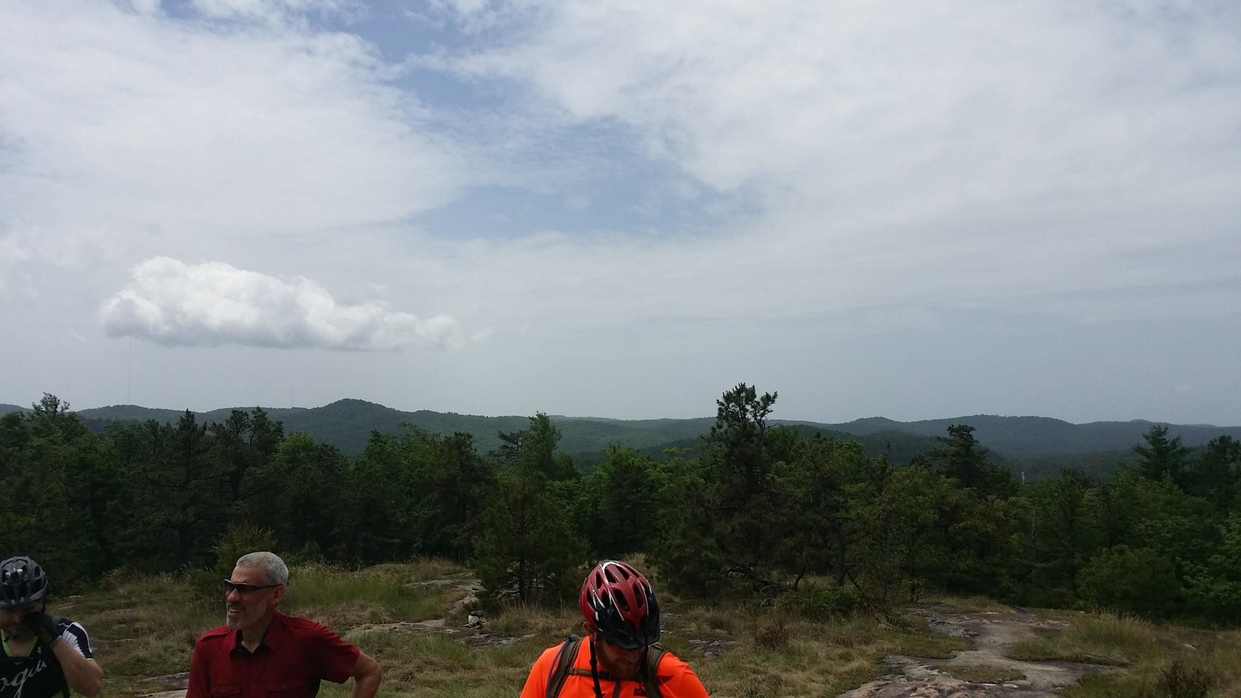 A scenic view from a rocky overlook featuring rolling hills and a forested landscape under a partly cloudy sky. Two individuals are visible in the foreground, one wearing a red shirt and the other in a bright orange shirt with a bicycle helmet. DuPont State Forest mountain bike trail.