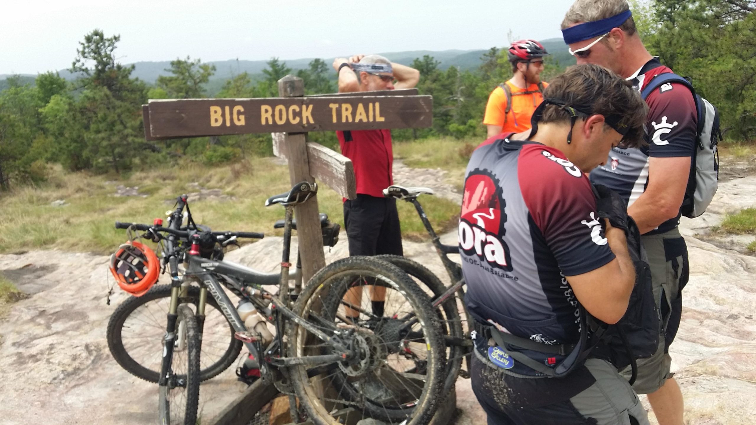 A group of mountain bikers at the Big Rock Trail sign, preparing for their ride. Two cyclists are adjusting their gear while another stands in the background, and several bikes with gear are parked nearby. The landscape features green trees and a rocky terrain. DuPont State Recreational Forest mountain bike trail.