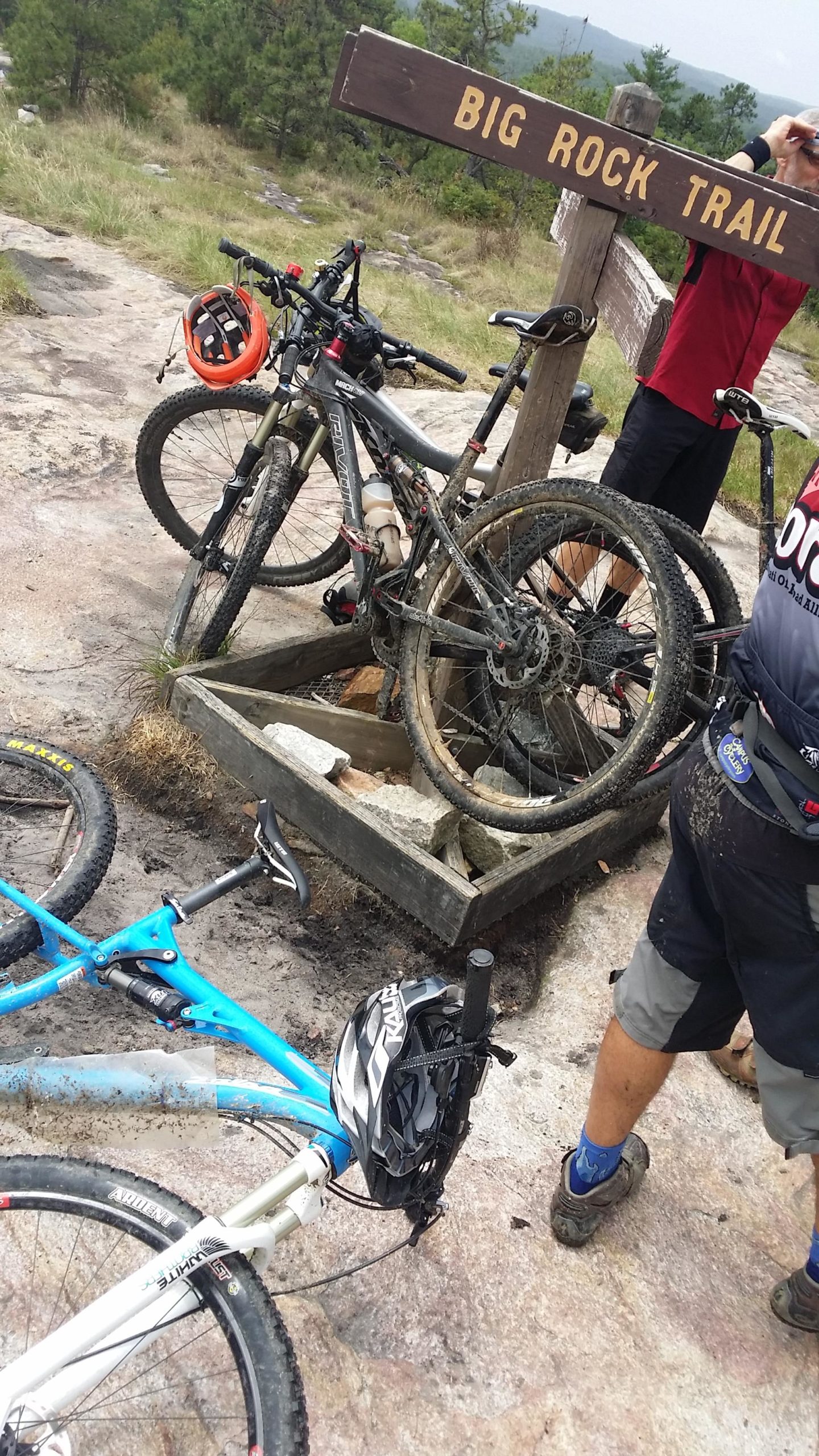 A mountain biking scene featuring two bicycles, one blue and one gray, resting near a wooden sign that reads "BIG ROCK TRAIL." The area shows rocky terrain and greenery in the background. A person in a red shirt is adjusting their gear nearby, while another cyclist is in shorts, wearing a helmet attached to their bike. The bicycles are muddy, indicating recent use on trails. DuPont State Forest mountain bike trail.