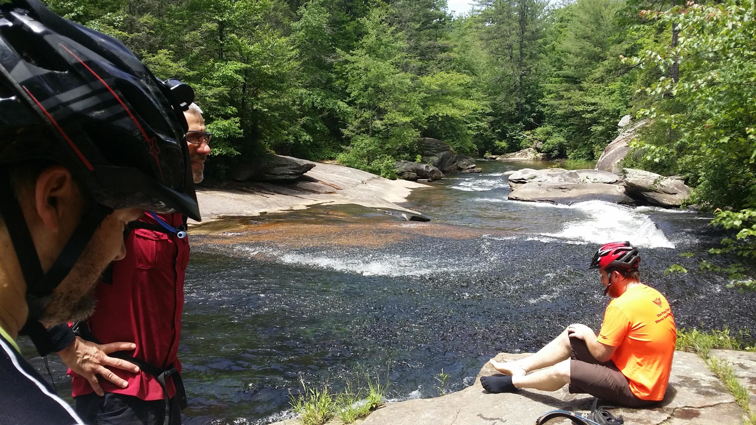 Three mountain bikers are enjoying a scenic riverside break. Two men are standing by the water, one wearing a black helmet and the other in a red shirt, as they gaze at the flowing river. Another man sits on a rock by the water, barefoot and wearing an orange shirt, with his legs dipped into the river. The lush greenery and rocky landscape create a peaceful outdoor setting. DuPont State Forest mountain bike trail.