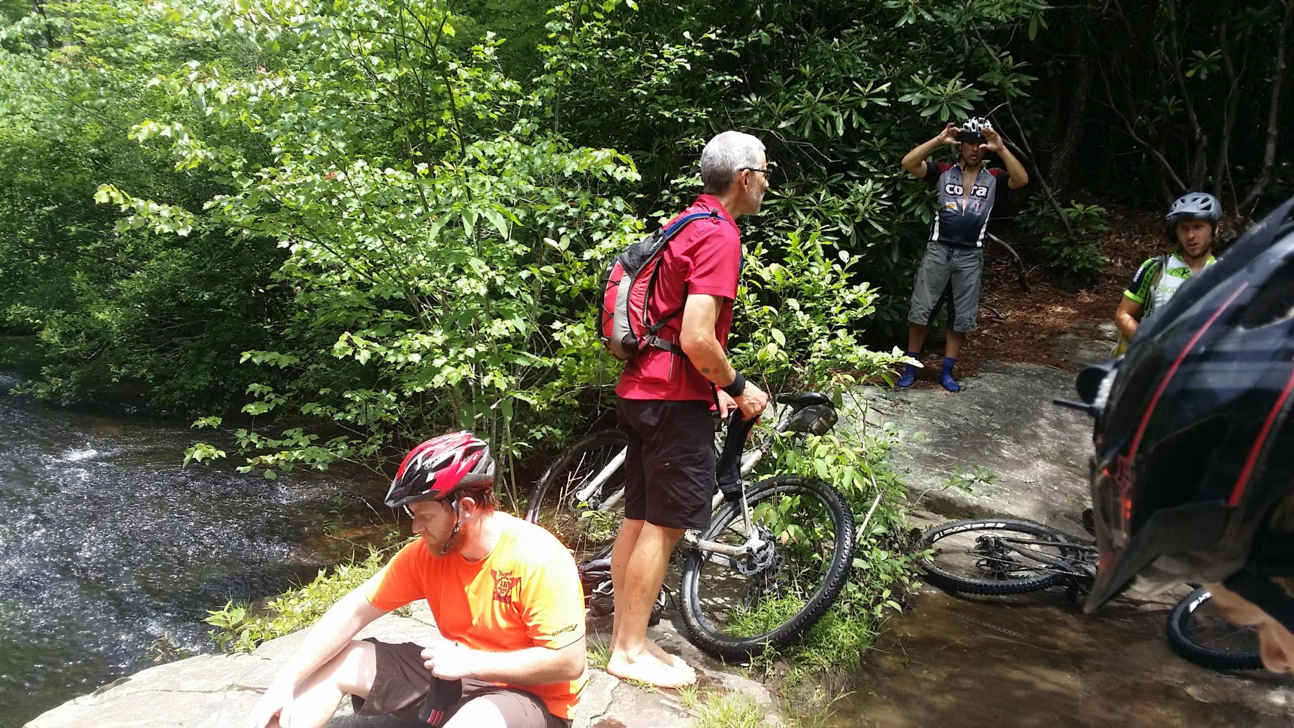 A group of mountain bikers taking a break by a flowing stream in a lush, green forest. One cyclist, wearing an orange shirt and a helmet, is sitting on a rock by the water, while another, dressed in a red shirt, is standing with a bike. Two other bikers are in the background, one adjusting his helmet and the other looking on. The scene captures a sunny day, with vibrant foliage surrounding the bikers. DuPont State Recreational Forest mountain bike trail.