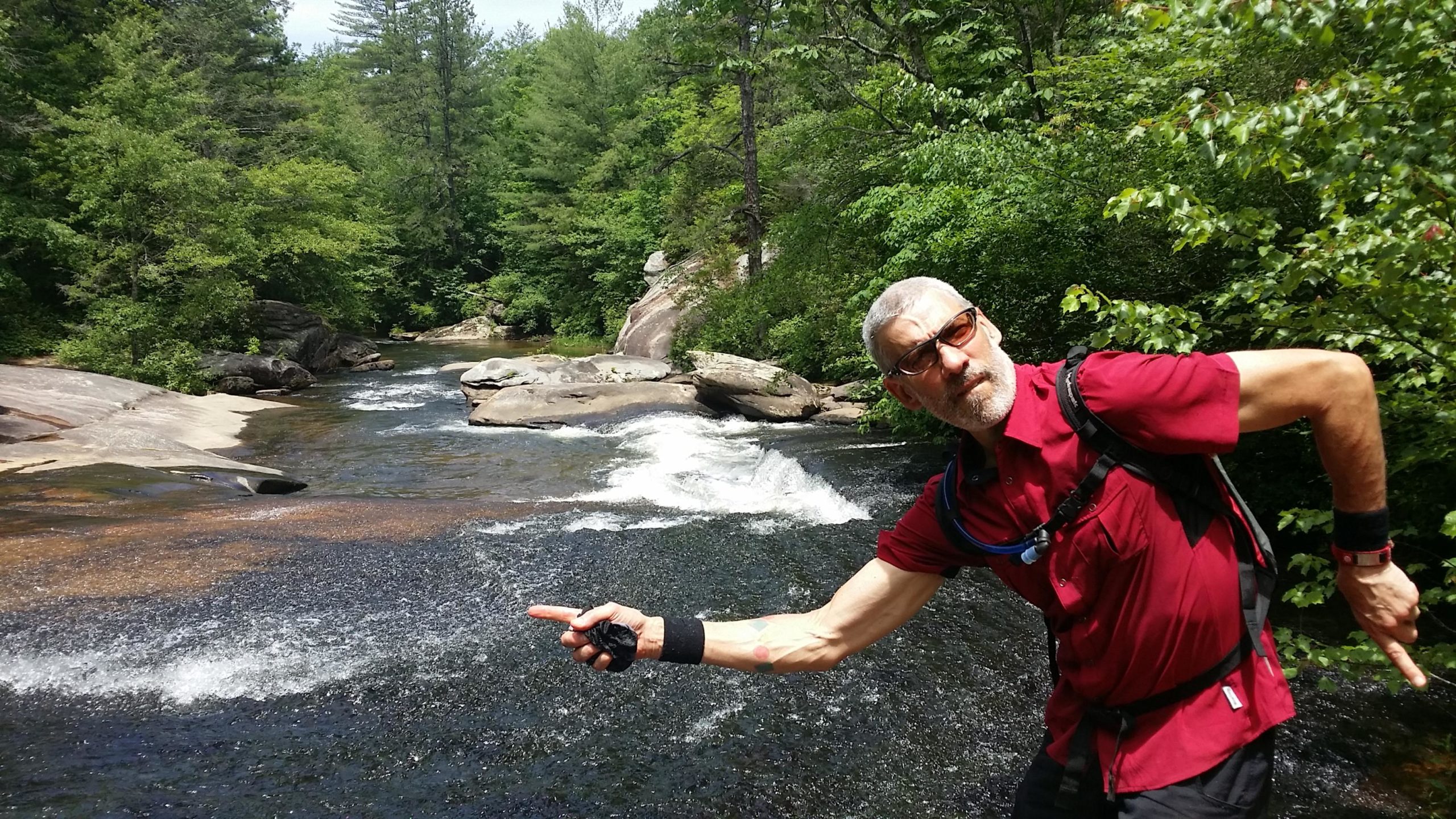 A man in a red shirt and sunglasses stands by a flowing river, pointing to his left. He is surrounded by lush greenery and rocky terrain, with water cascading over stones in the background under a clear sky. DuPont State Recreational Forest mountain bike trail.